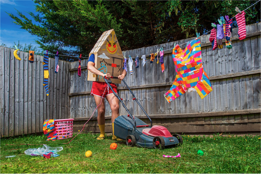 A man in bright red shorts and long yellow socks with a cardboard house covering his head and torso holds a lawn mower in a back garden next to a washing line with several brightly coloured garments hanging on it.