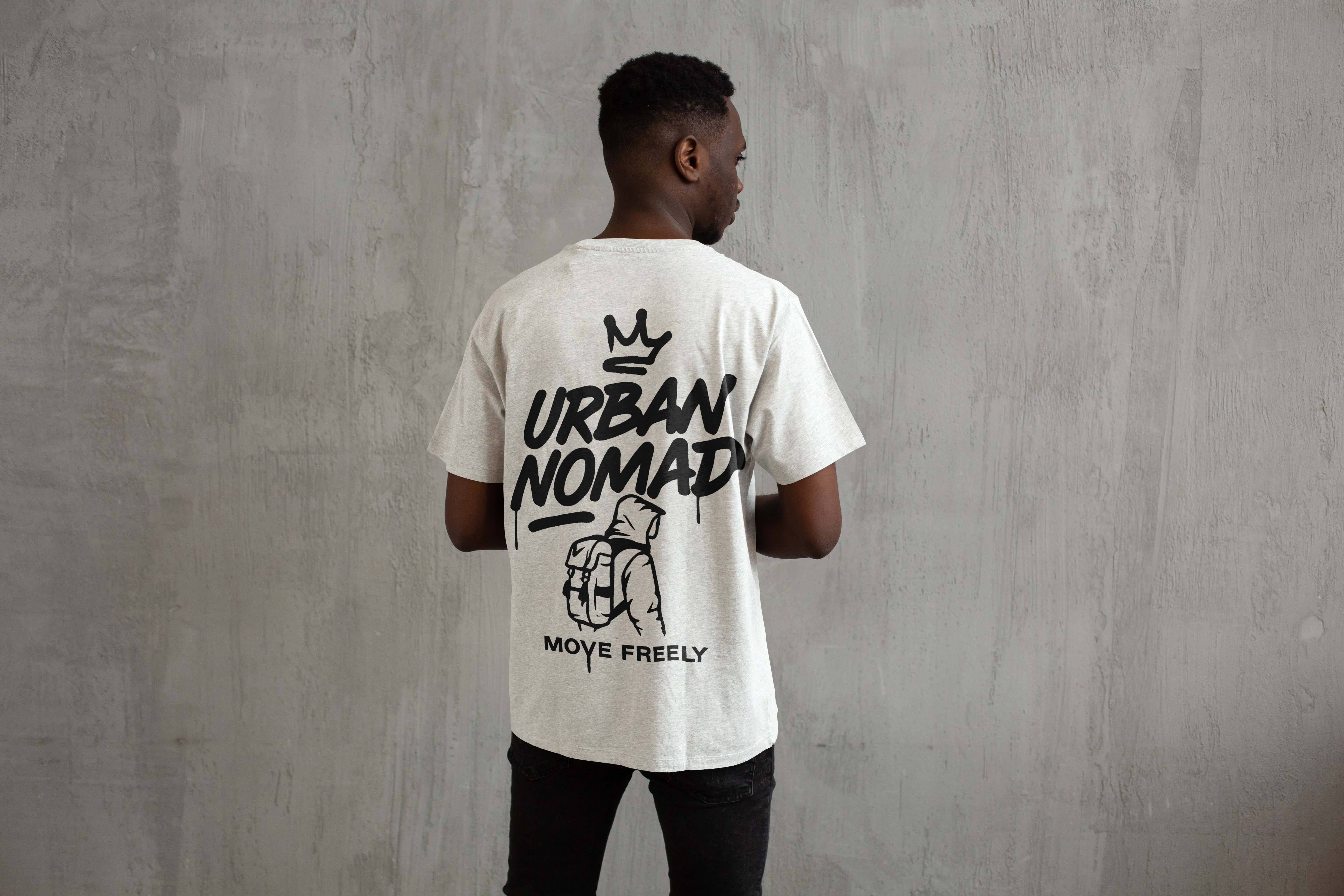 Man facing away wearing white t-shirt with "URBAN NOMAD" text, crown logo, and "MOVE FREELY" slogan, against concrete wall.