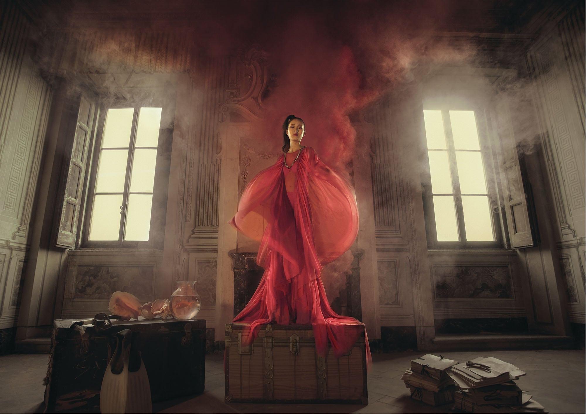 Lady in red dress stands on a chest with smoke coming out from behind her. There is a luggage trunk to her right and a pile of documents to her left.