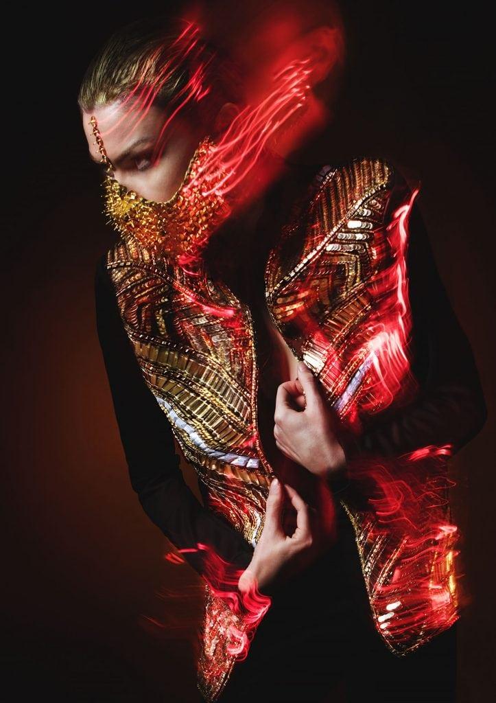 Person wearing a sequinned jacket and a gold face mask poses side-on.