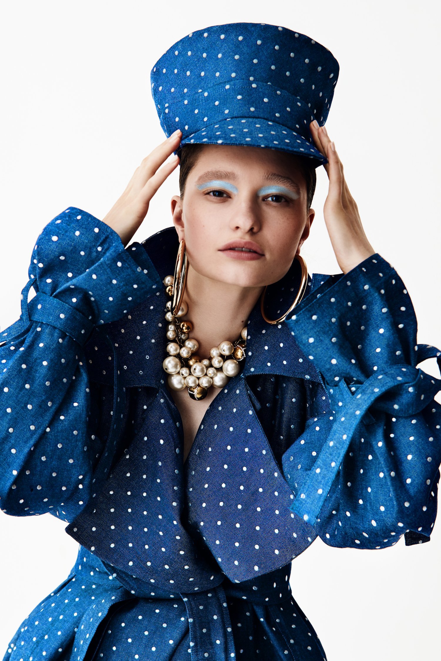 A model wearing a blue spotted dress and hat, with a silver-gold bubble neckpiece.