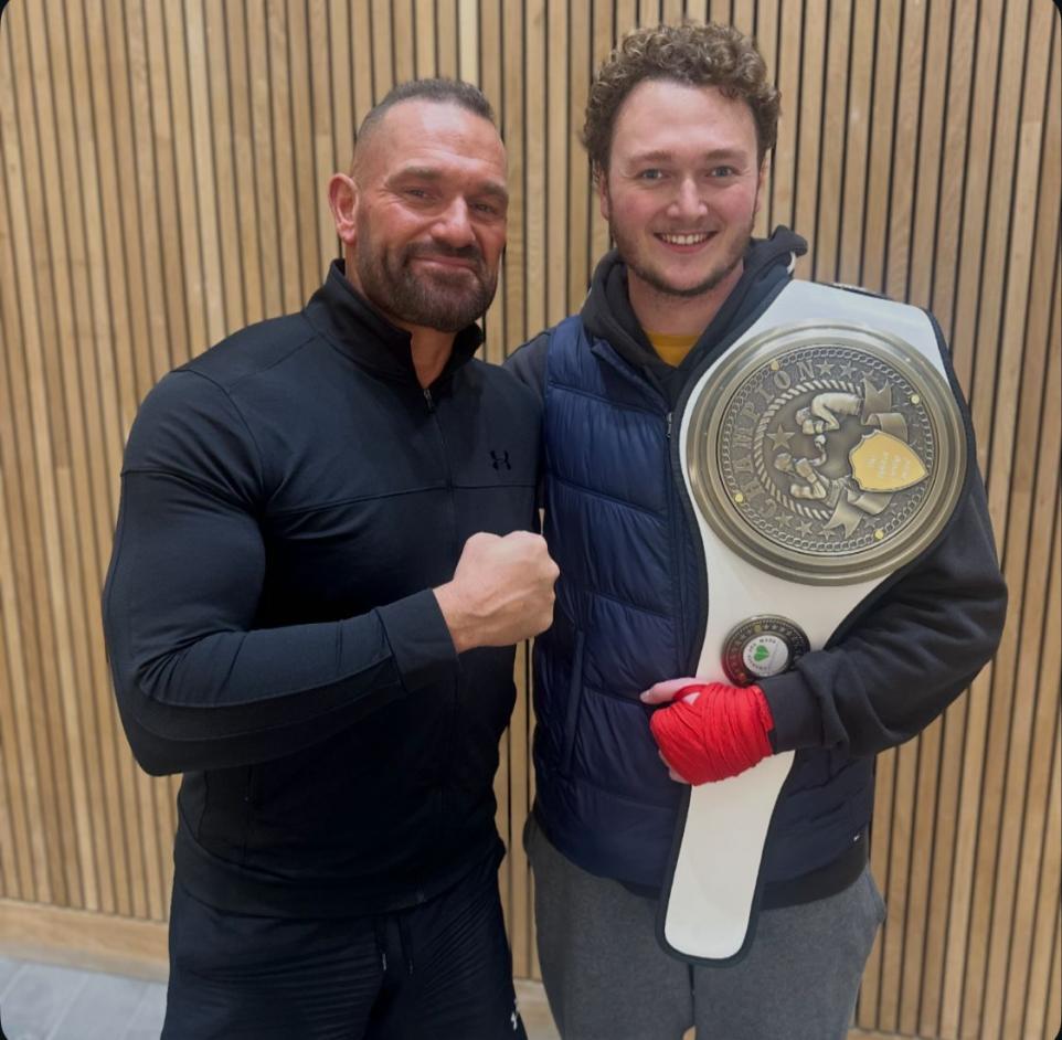 M Maguire and M Smith posing together, one holding a large championship belt. Man on left wears black jumper, man on right wears navy jacket.