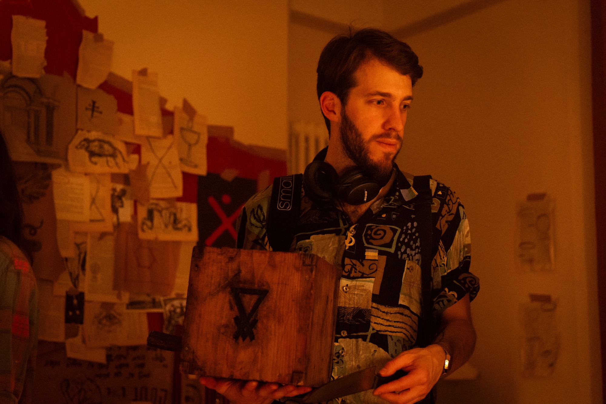 BA (Hons) Film Production graduate M R Lincoln holding a box on a film set. A wall was loads of papers and clippings tacked to it is in the background.