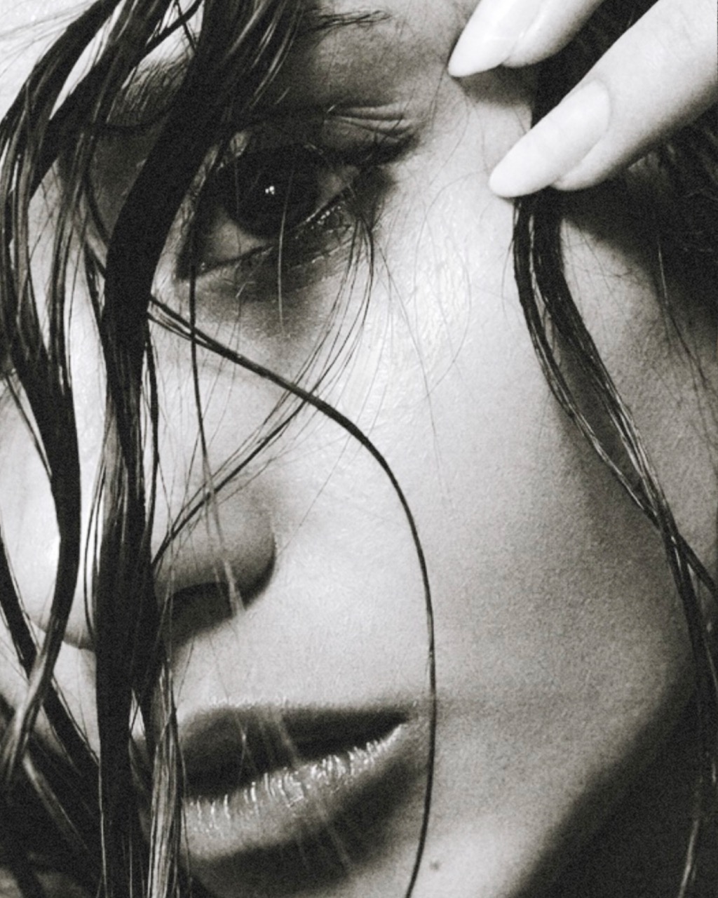 A close-up black and white image of a woman's face with wet hair partially covering her forehead. Her hand is placed near her hair, and the focus is on her eye and lips, exhibiting fine details of her skin texture.