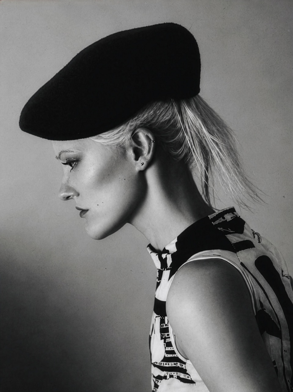 A woman with short blonde hair styled in a ponytail is posed in profile, wearing a black beret and a patterned sleeveless top. The image is in black and white, highlighting her facial features against a neutral background.