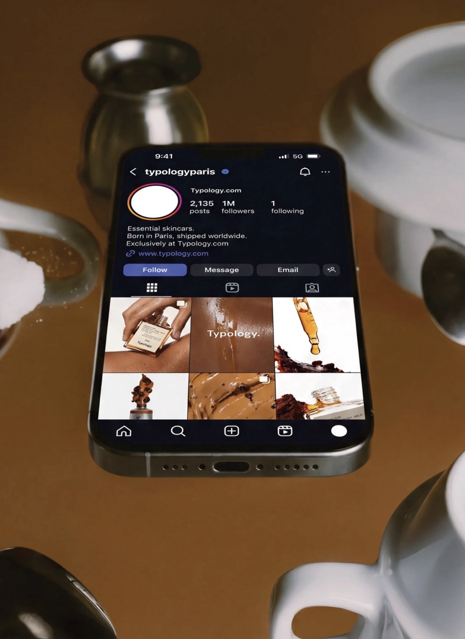 A smartphone displaying an Instagram profile for 'typologyeparis', featuring posts related to essential skincare. Surrounding the phone are silver teapots and cups on a brown surface.