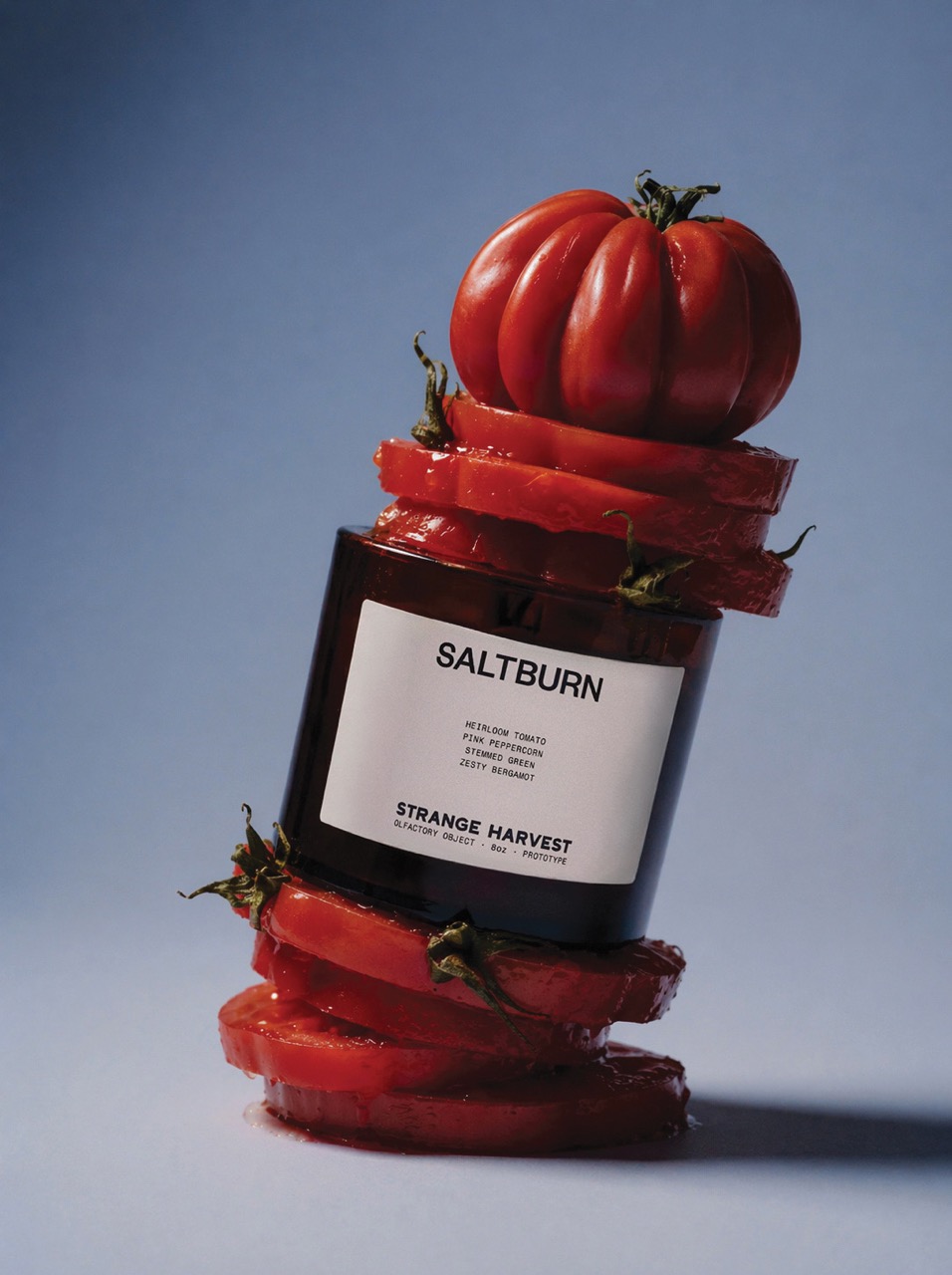 A brown glass jar labelled 'SALTBURN' with a white label sits among overlapping slices of red tomatoes. A whole red tomato topped with green leaves is placed above the jar against a soft blue background.