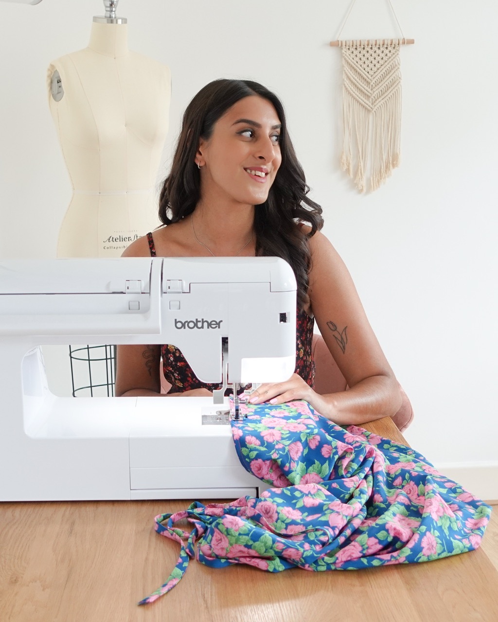 Tammy Johal sat at a wooden table sewing a floral dress with a machine, mannequin and dream catcher in the background.