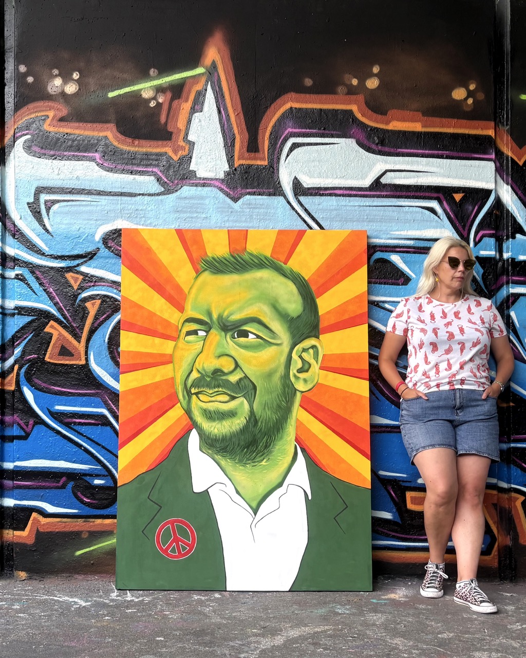 A woman in sunglasses stands beside a colourful portrait painting leaning against a graffiti-covered wall.