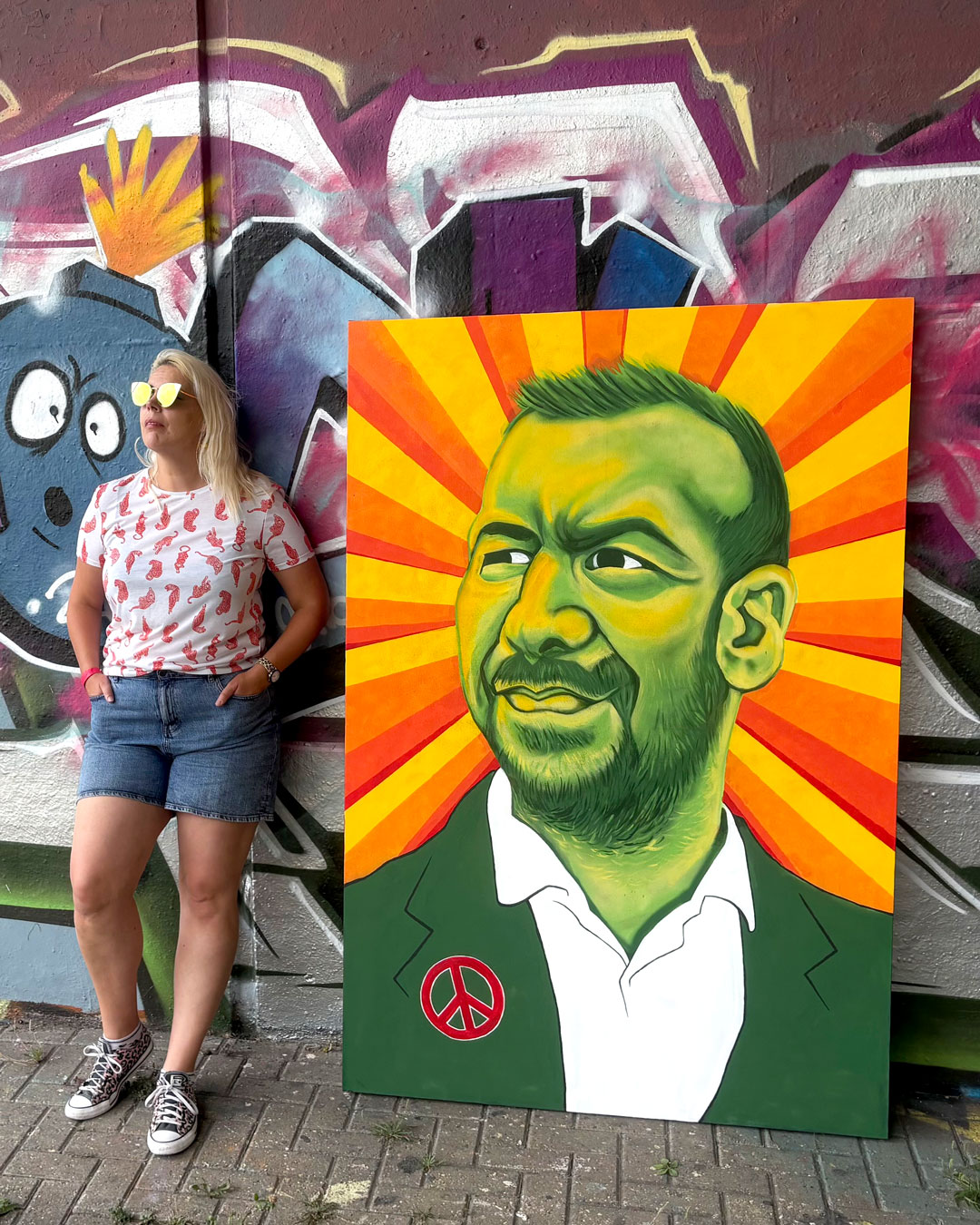 A woman in sunglasses and denim shorts stands beside a brightly coloured portrait painting leaning against a graffiti-covered wall.