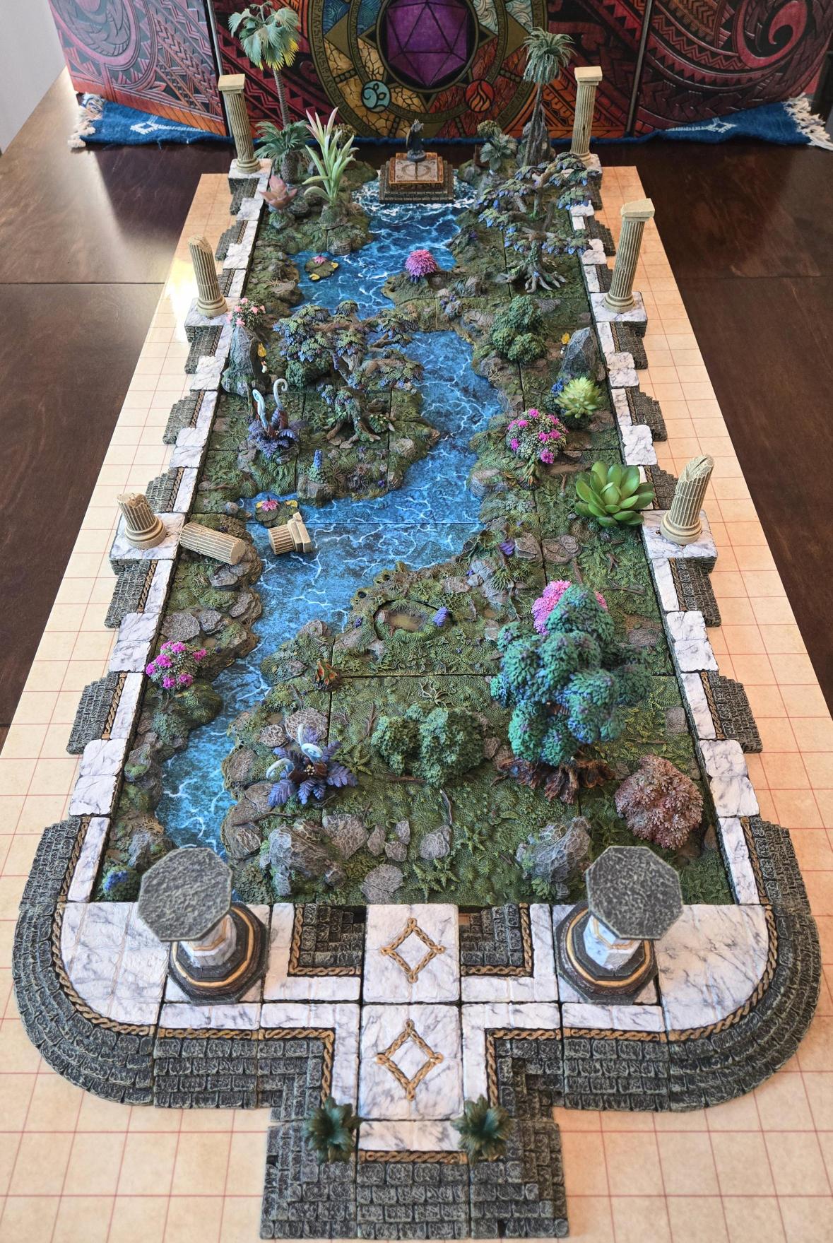 A rectangular miniature landscape model with a winding blue stream, rocks and plants, bordered by stone-like paths and columns on a tiled floor.