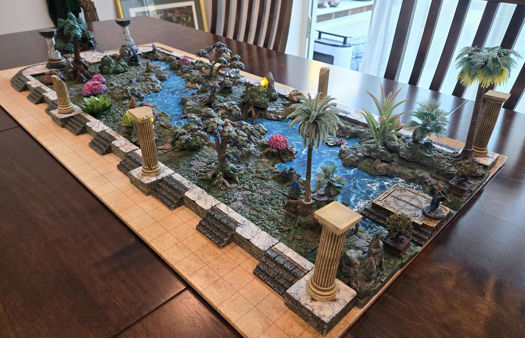 A detailed tabletop model of a rocky oasis with a blue waterway, palm trees and small stone columns, displayed on a wooden dining table.