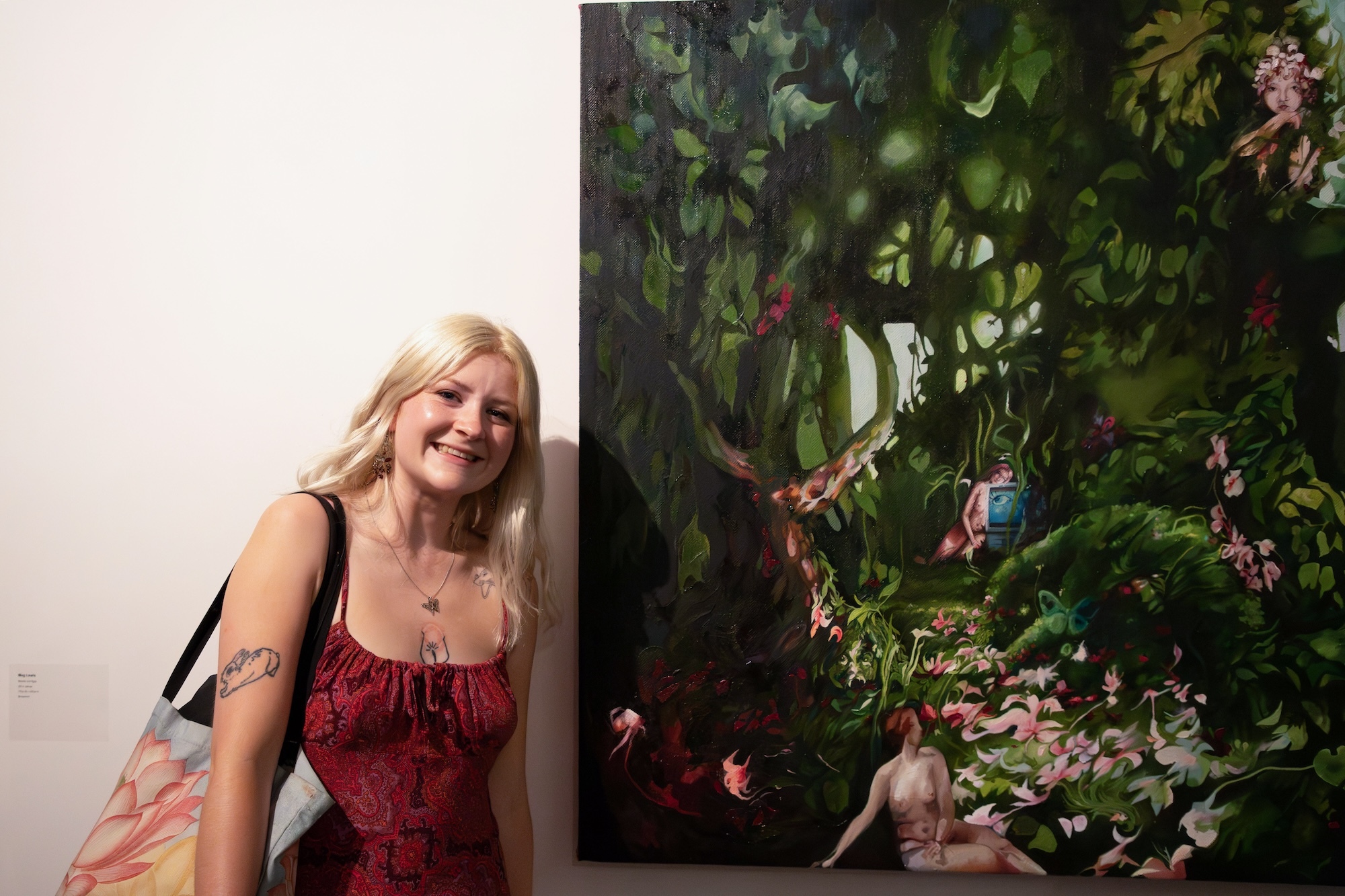 Artist M Lewis standing next to a canvas of a painting of a deep green forest scene with two nude figures visible.