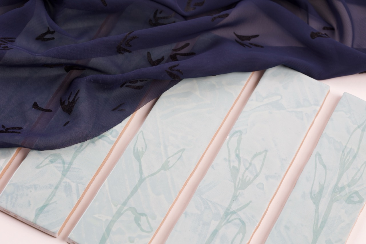 A collection of four rectangular tiles with a light blue floral pattern on a white background, partially covered by a sheer navy fabric with black floral designs. The tiles are arranged neatly against a light surface.