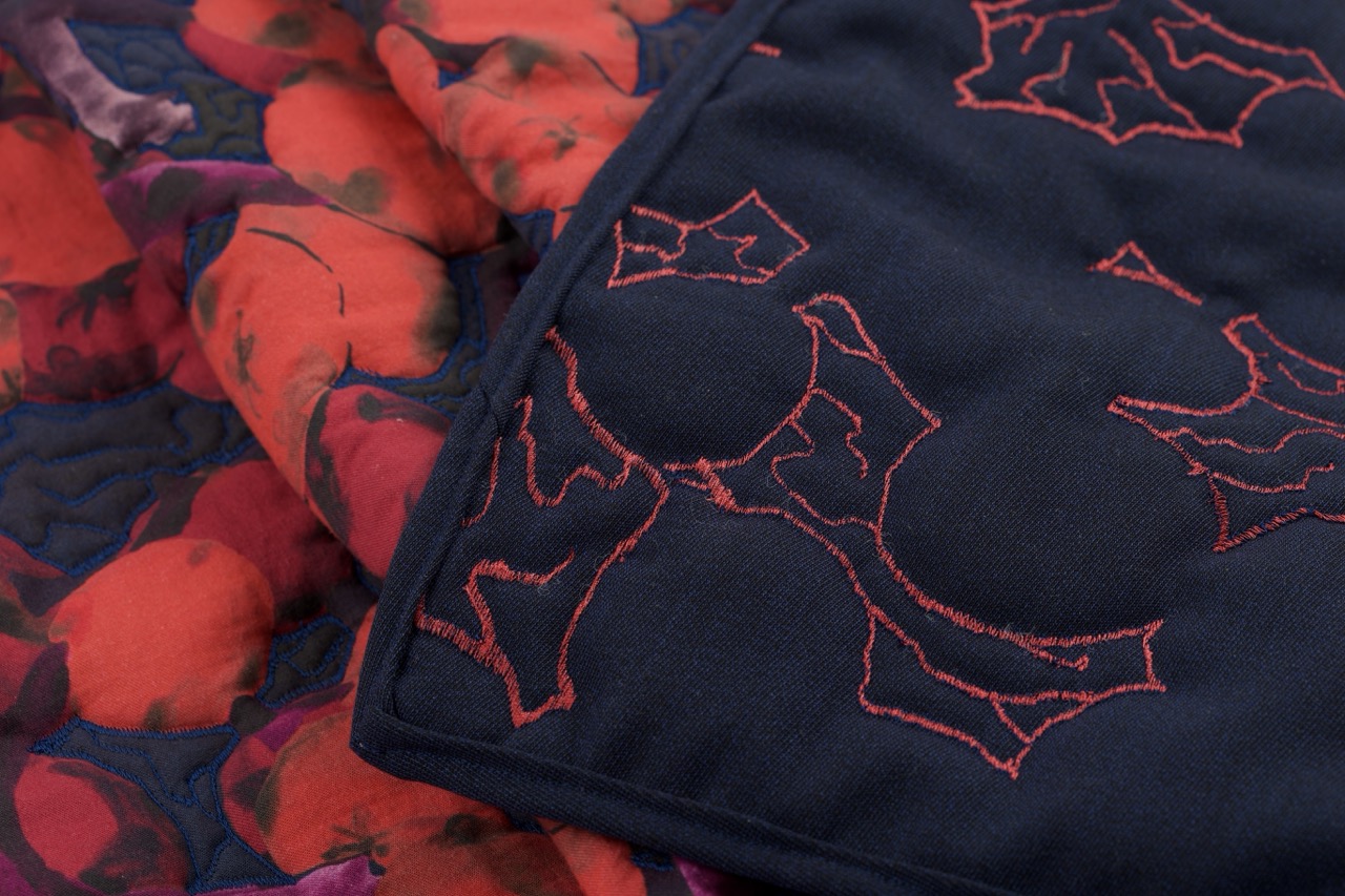 A close-up view of a patterned fabric featuring vibrant floral designs in shades of red and blue, with a dark section displaying intricate red embroidery of holly leaves and berries.