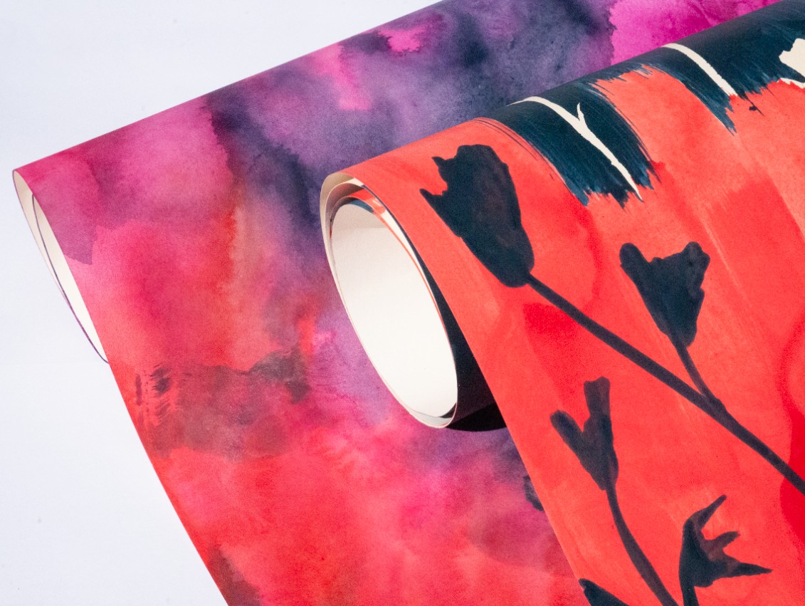 Two rolls of decorative wrapping paper. One features a vibrant pink and purple watercolour design, while the other displays an orange background with black floral silhouettes. The rolls are partially unrolled, showcasing their vivid patterns.