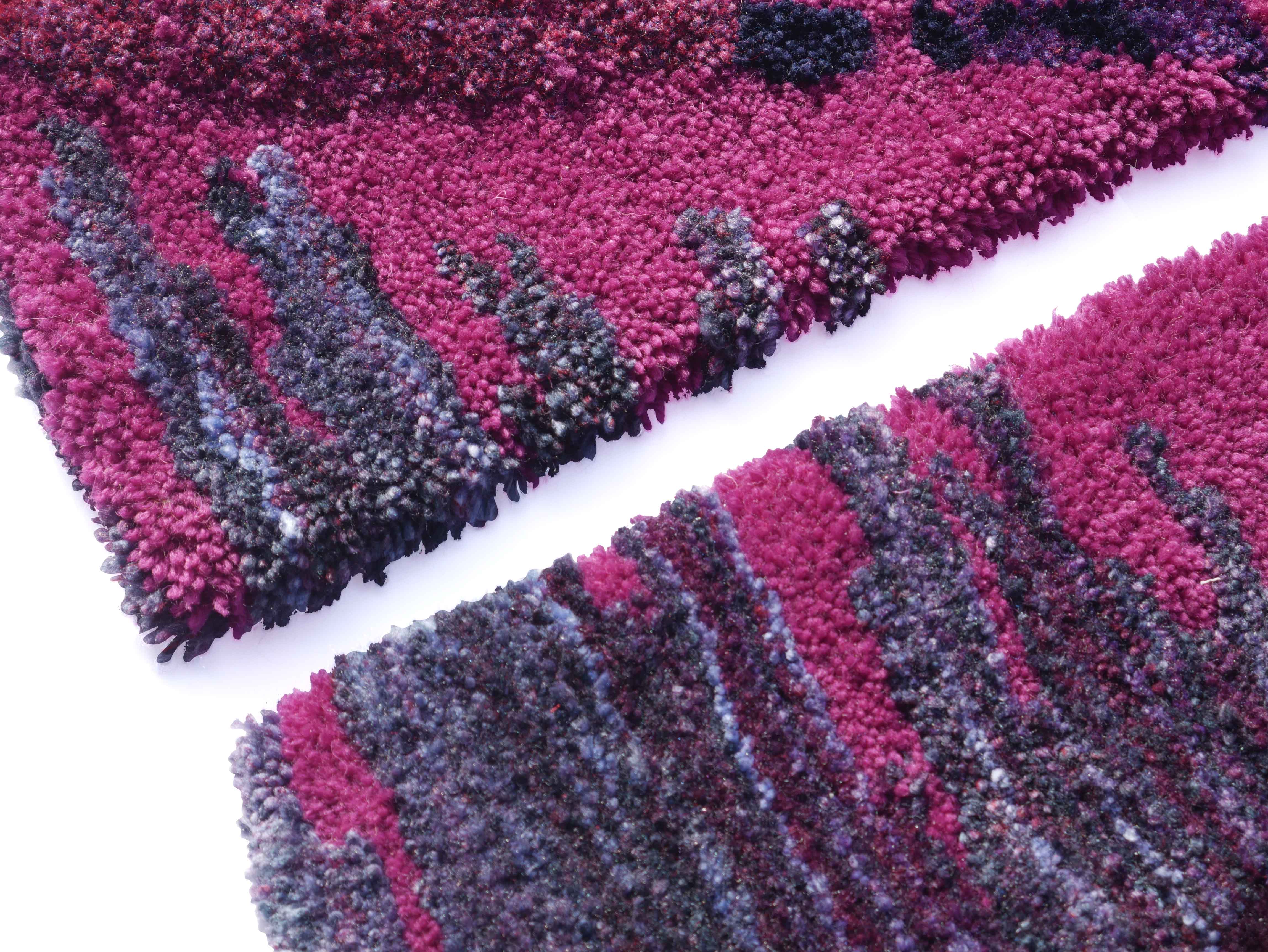 A close-up view of two textured rugs in varying shades of pink, purple, and grey. The rugs display a wavy pattern with multicoloured tufts, highlighting the rich texture and artistic design.