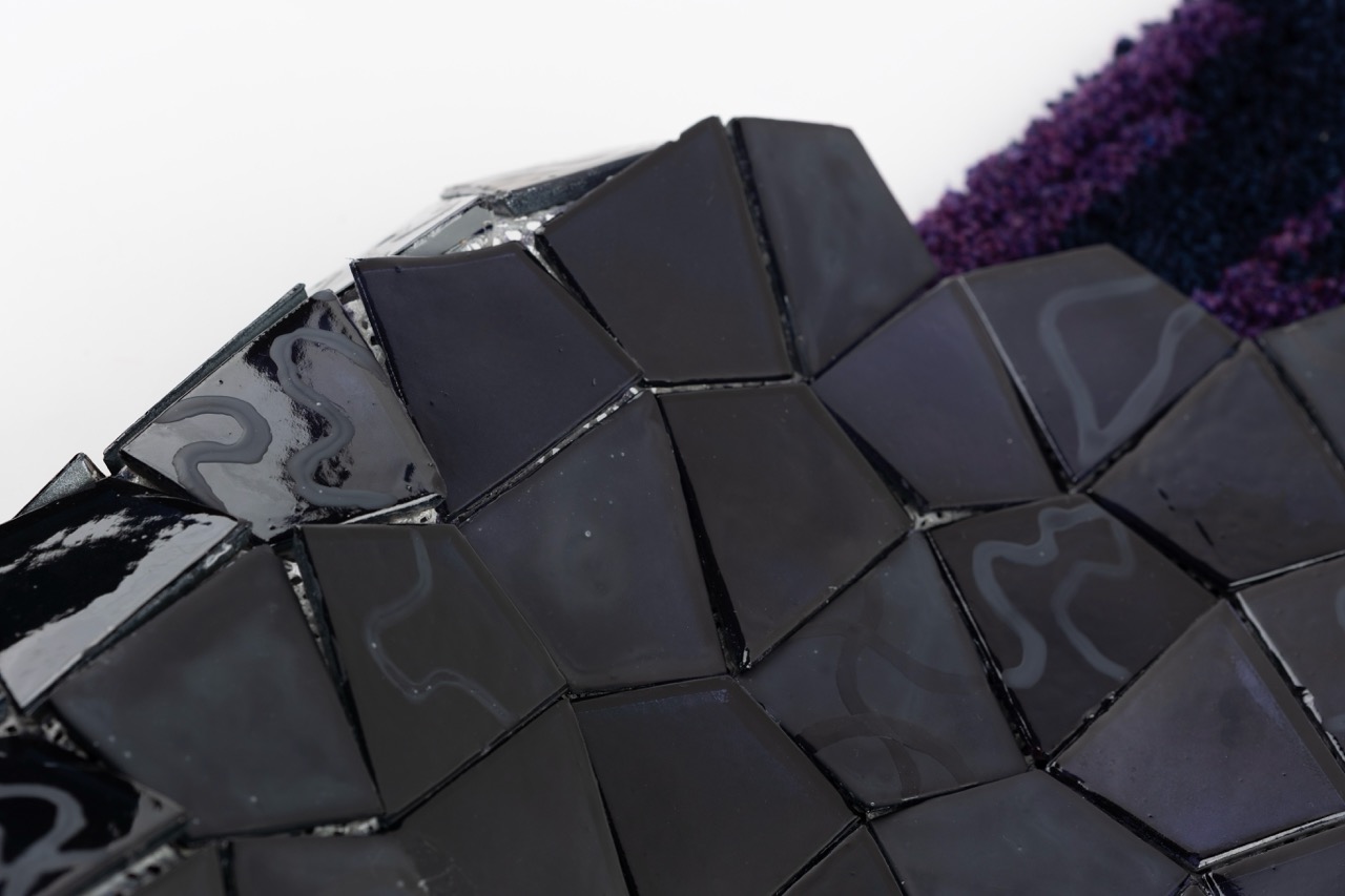 An abstract surface composed of geometric black tiles arranged in a mosaic pattern, reflecting light in various directions. The background features a section of purple fabric with a textured appearance.