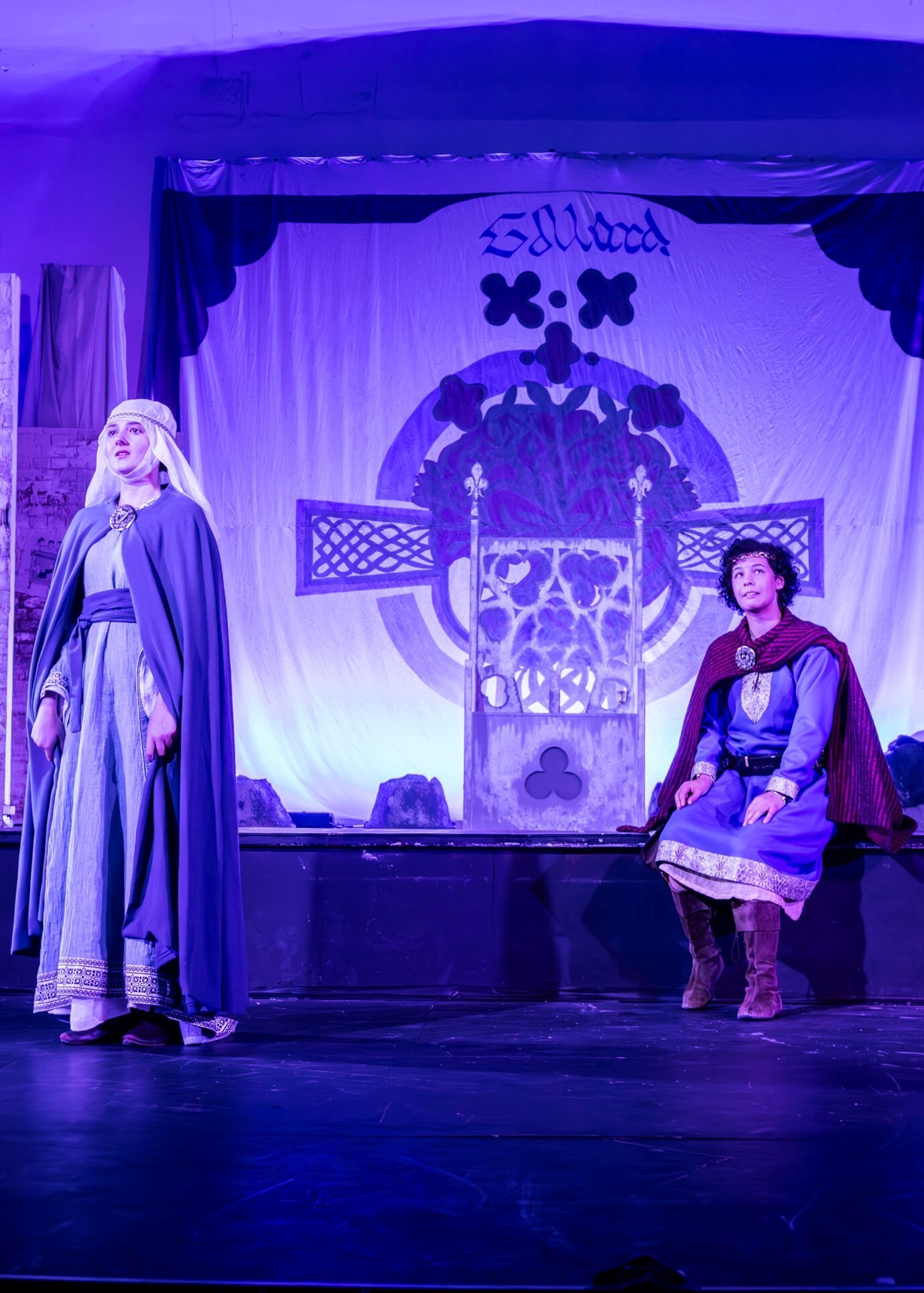 Two actors in medieval costumes on stage with purple lighting. Large skull backdrop with crossbones. Woman in grey robes left, man in burgundy tunic seated right.