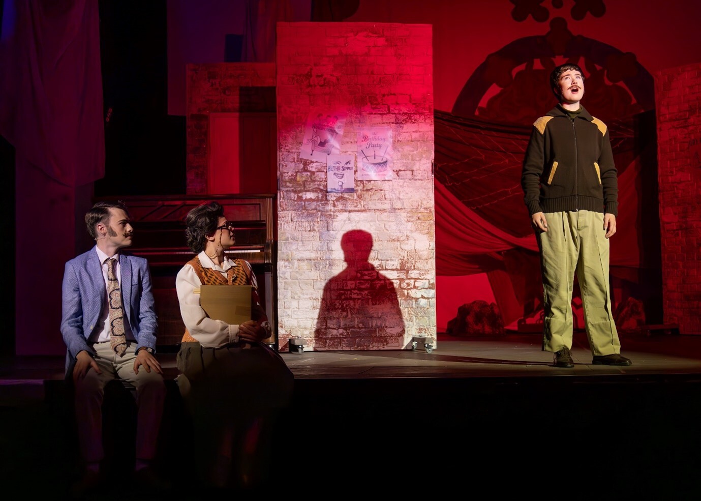 Theatre stage with three actors under red lighting. Stone wall backdrop with brick archway. Man in blue suit seated left, woman in brown vest centre, man in green jumper standing right.