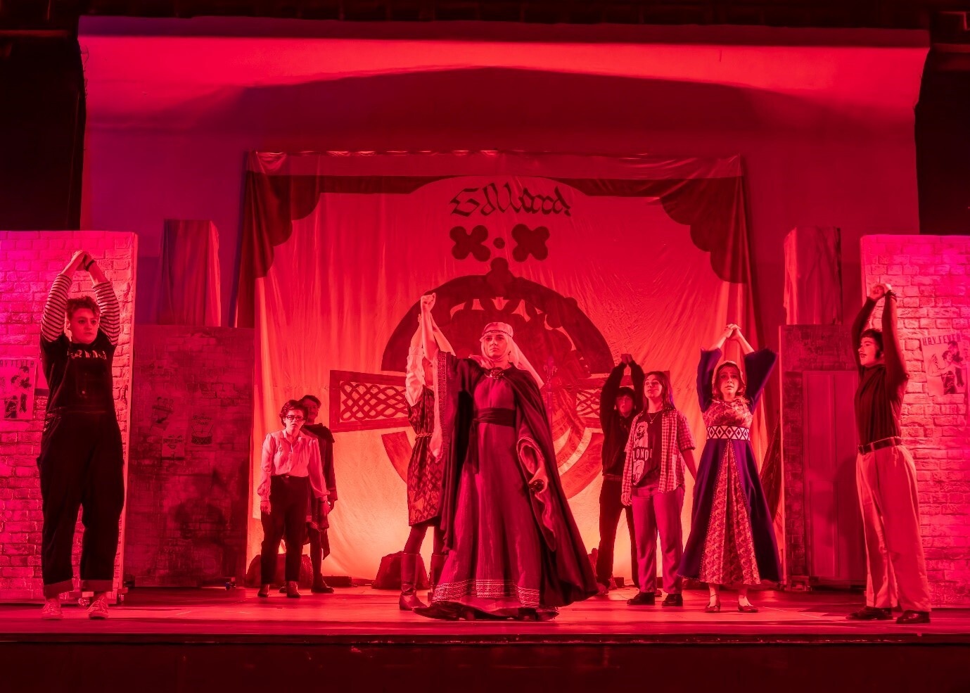Theatre stage bathed in red lighting with performers in period costumes arranged across ornate set with arched doorway and columns.