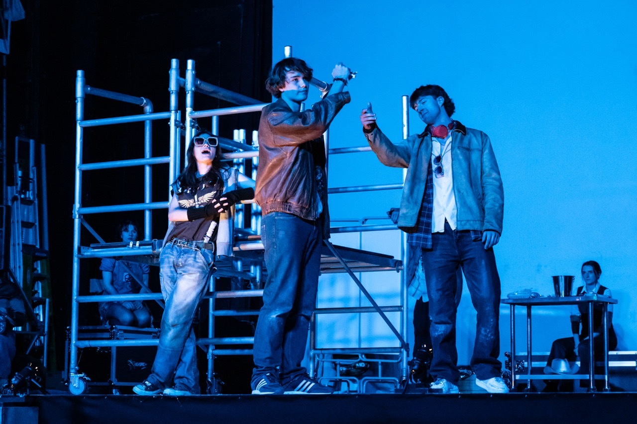 Three performers on stage with metal scaffolding and blue lighting, one holding microphone, another with drink, industrial set design.