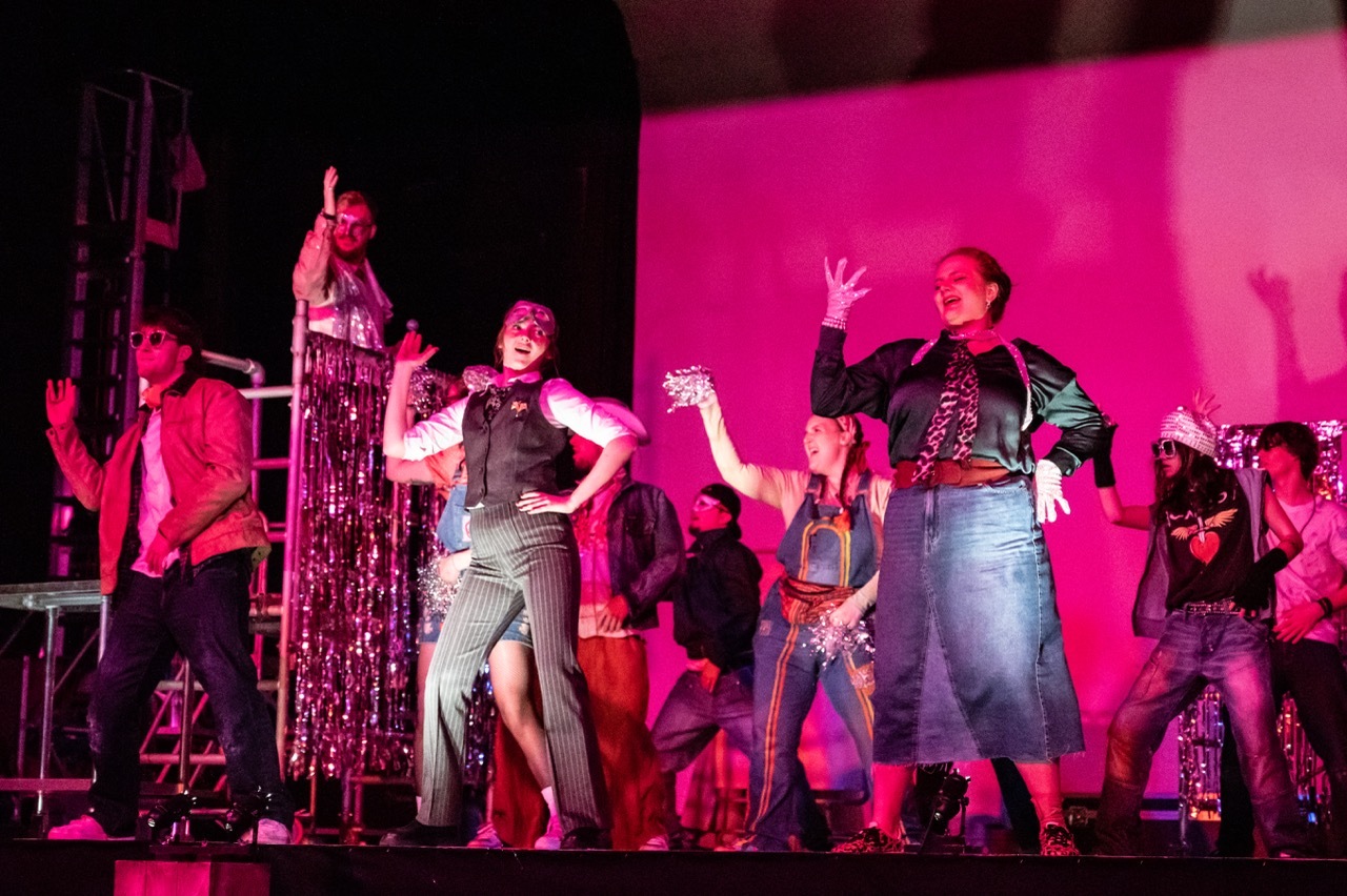 Theatre performers in period costumes on stage with bright pink lighting and dark background.