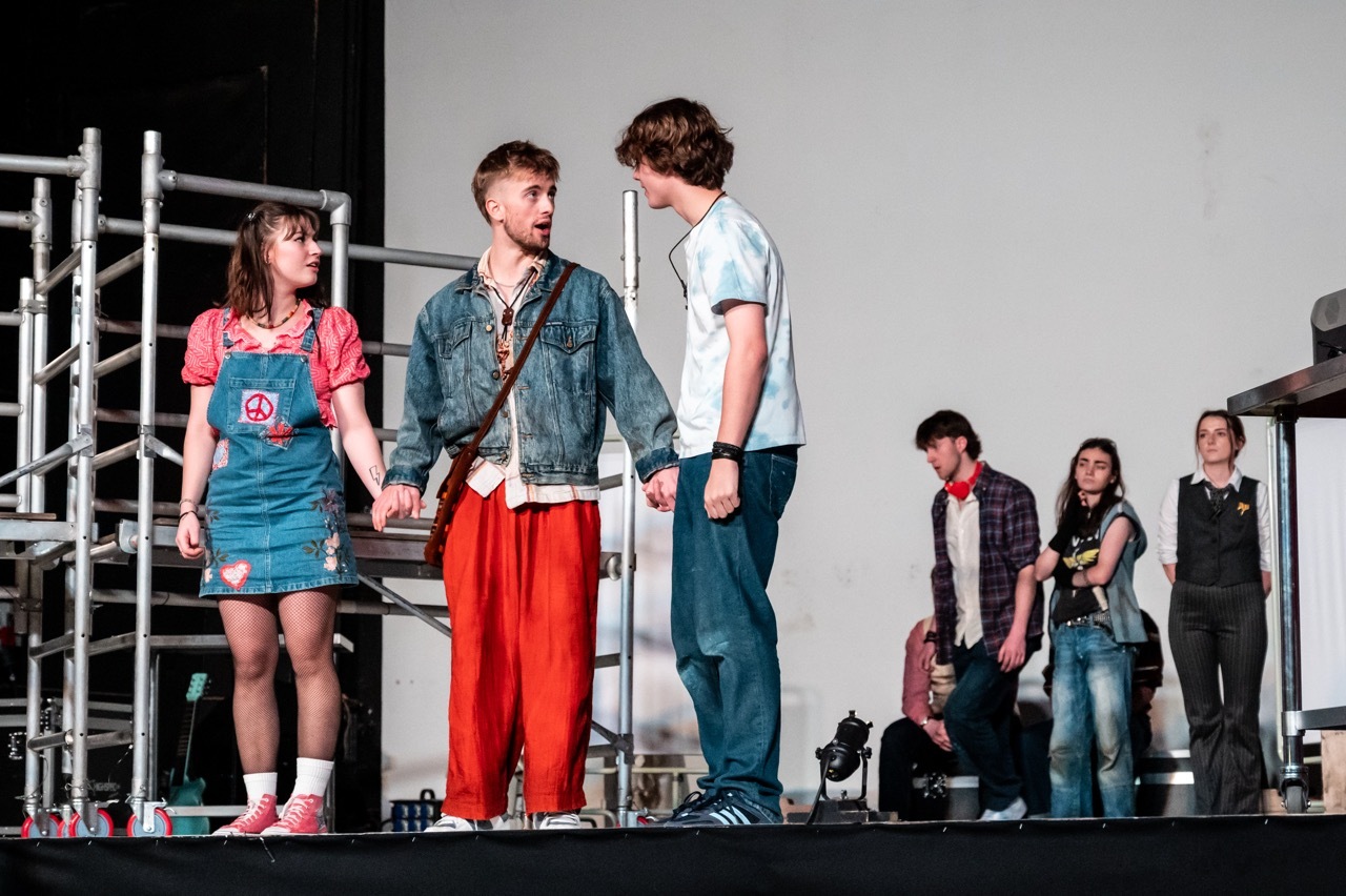 Three young actors on stage performing a scene, with additional cast members visible in background on metal scaffolding set.