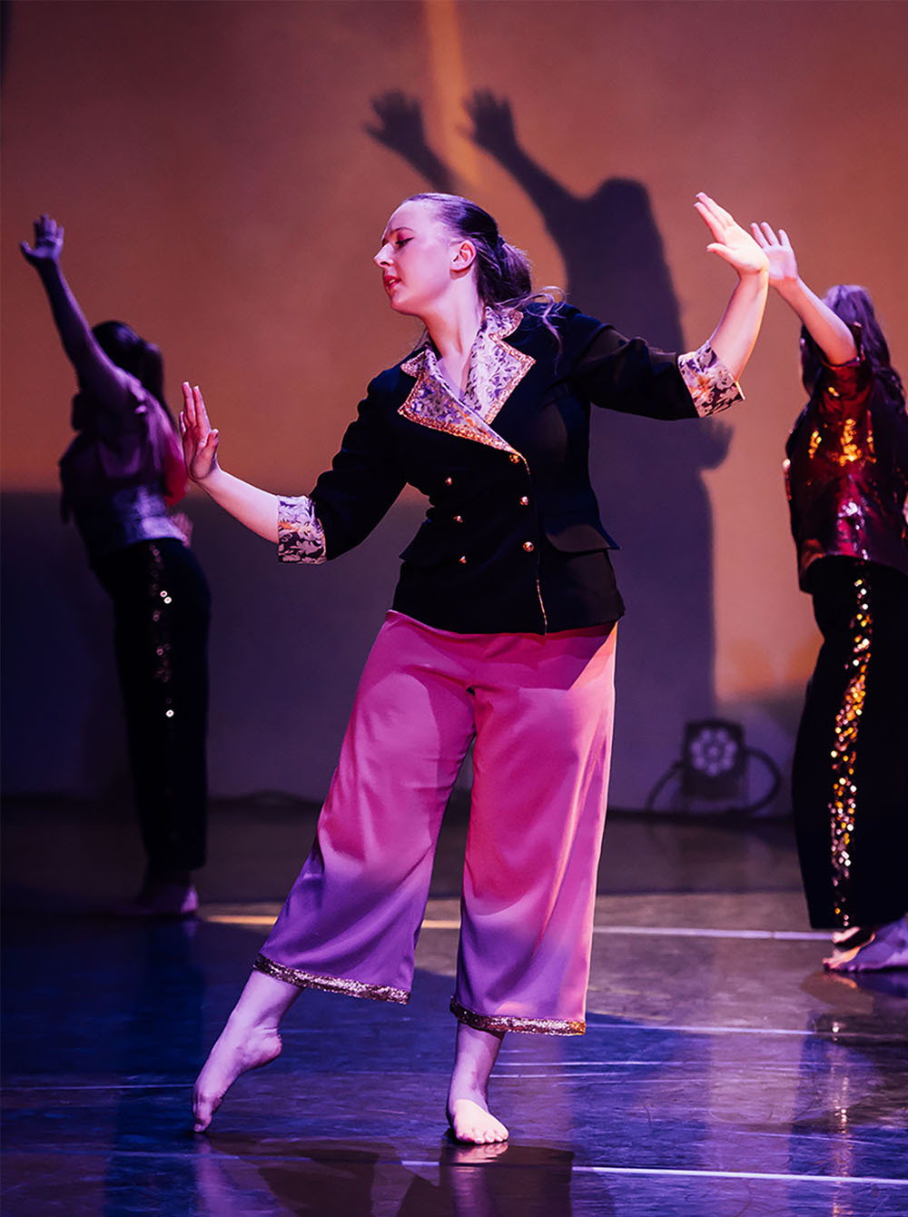 Female dancer in black embroidered top and pink trousers performs on stage with arms raised, other dancers visible in background.