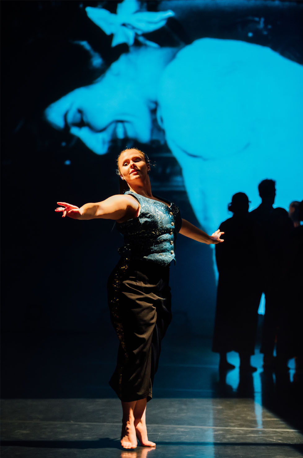 Female dancer in grey top and dark skirt on stage with extended arms. Large blue projection of face behind her, silhouettes visible.