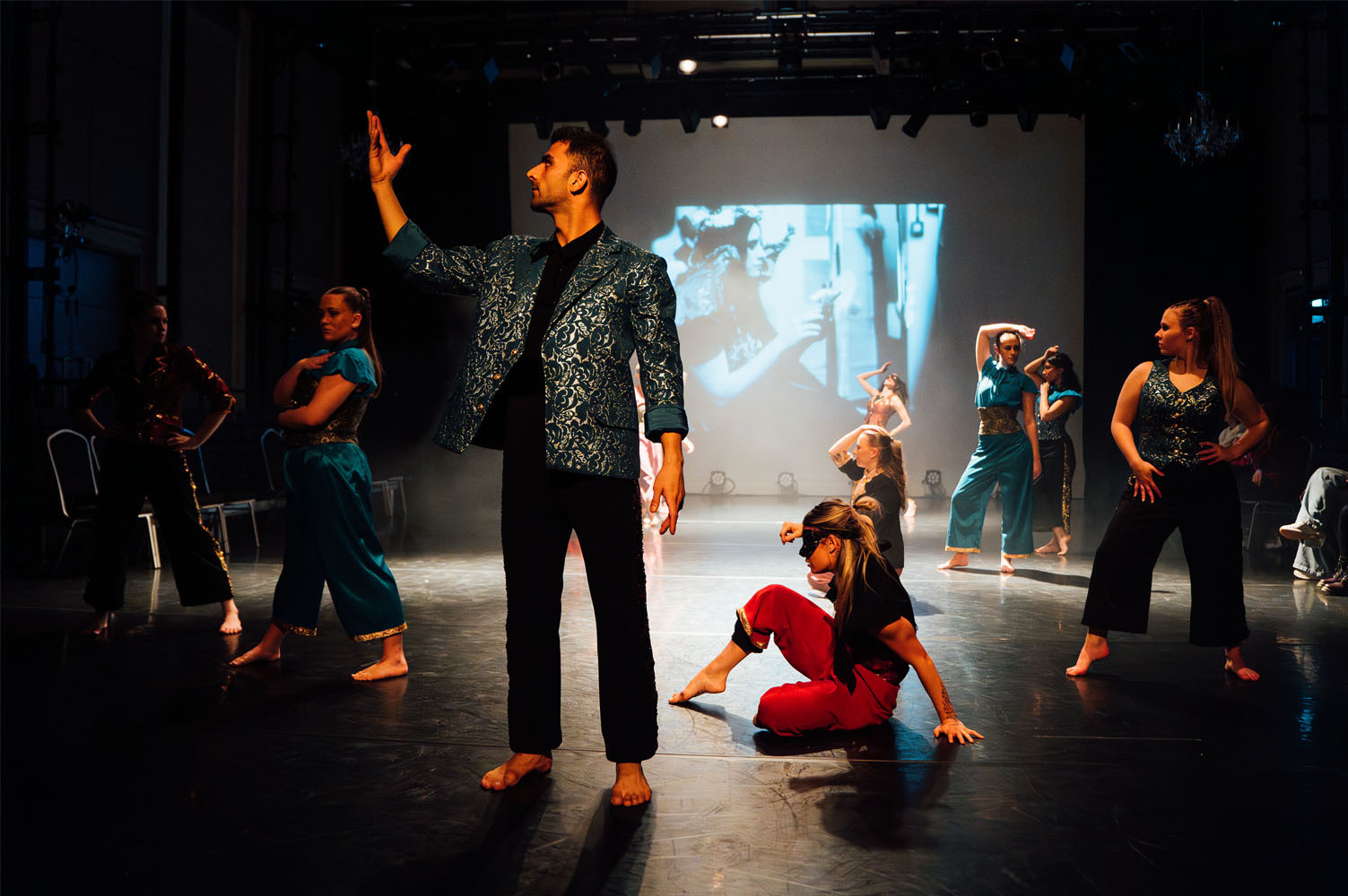 Theatre performance with actors in dark clothing on stage, one person kneeling in red, projection screen visible in background.