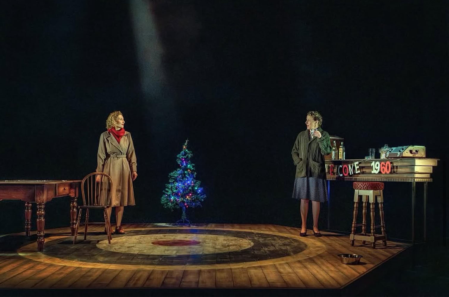 Two actors on wooden stage with small blue Christmas tree between them. Woman in beige coat on left, man at desk on right. Dark background with warm stage lighting on circular rug.