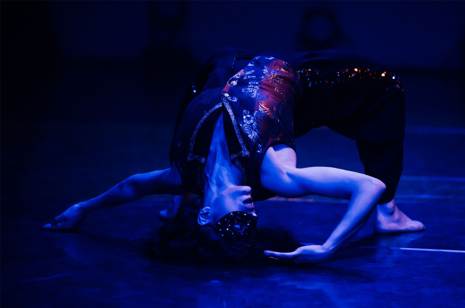 Performer in ornate mask crouched on stage floor under blue lighting, wearing dark costume with decorative headpiece.