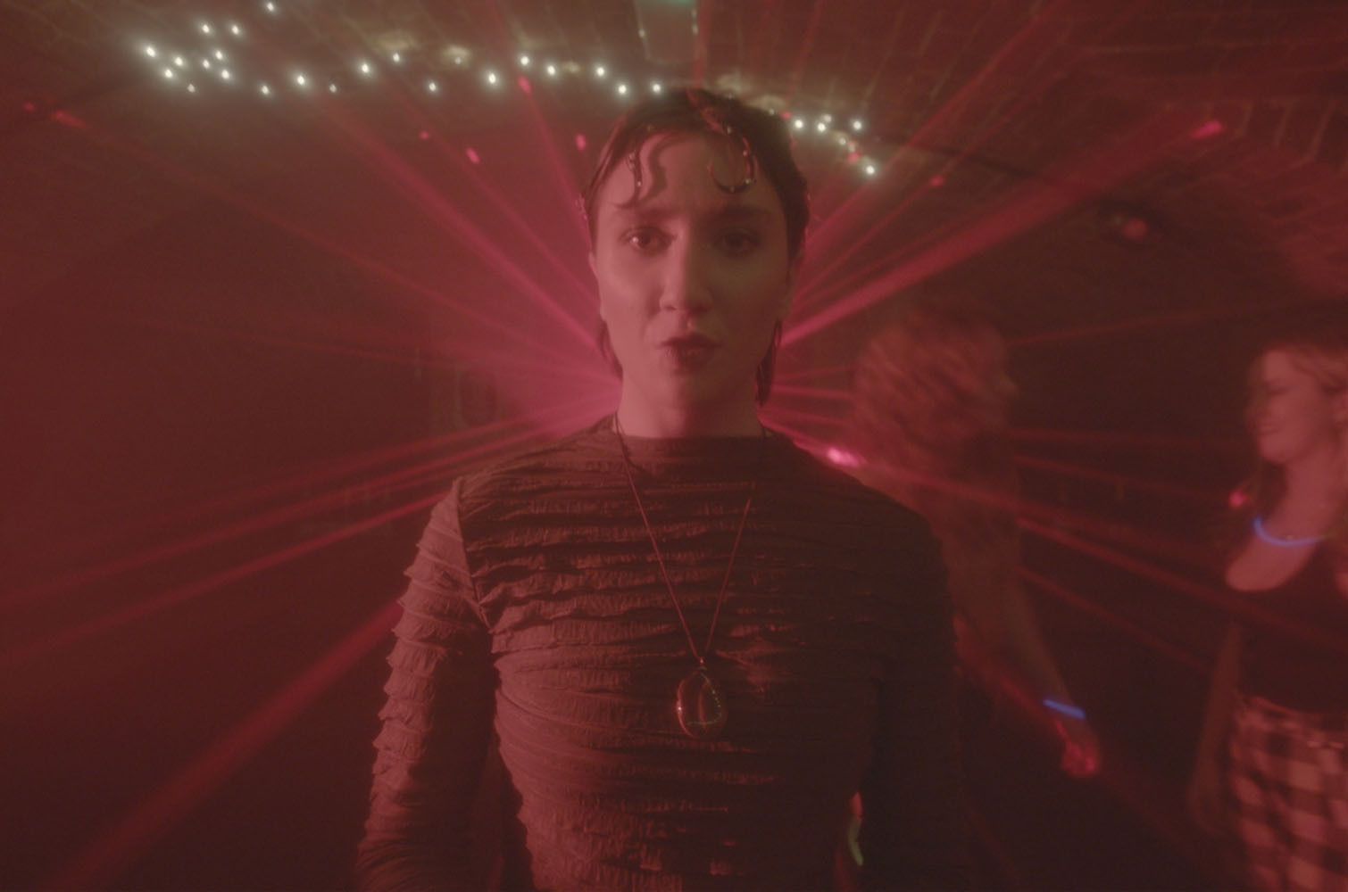 Man in striped jumper under red lighting with string of white bulbs overhead, blurred figures in background.