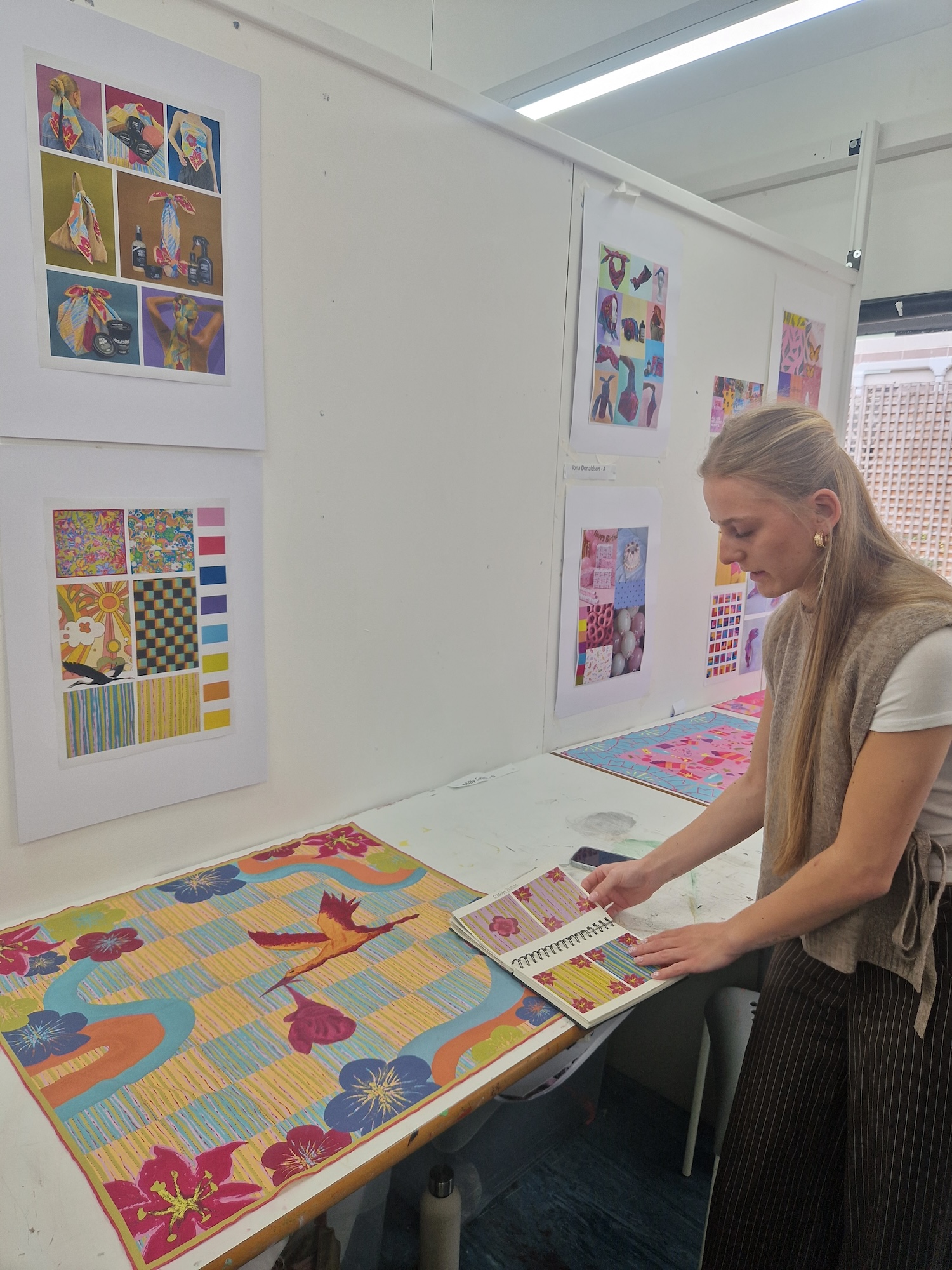 BA (Hons) Textiles Design student M Cullingford at a studio desk displaying bright, colourful floral textile designs.