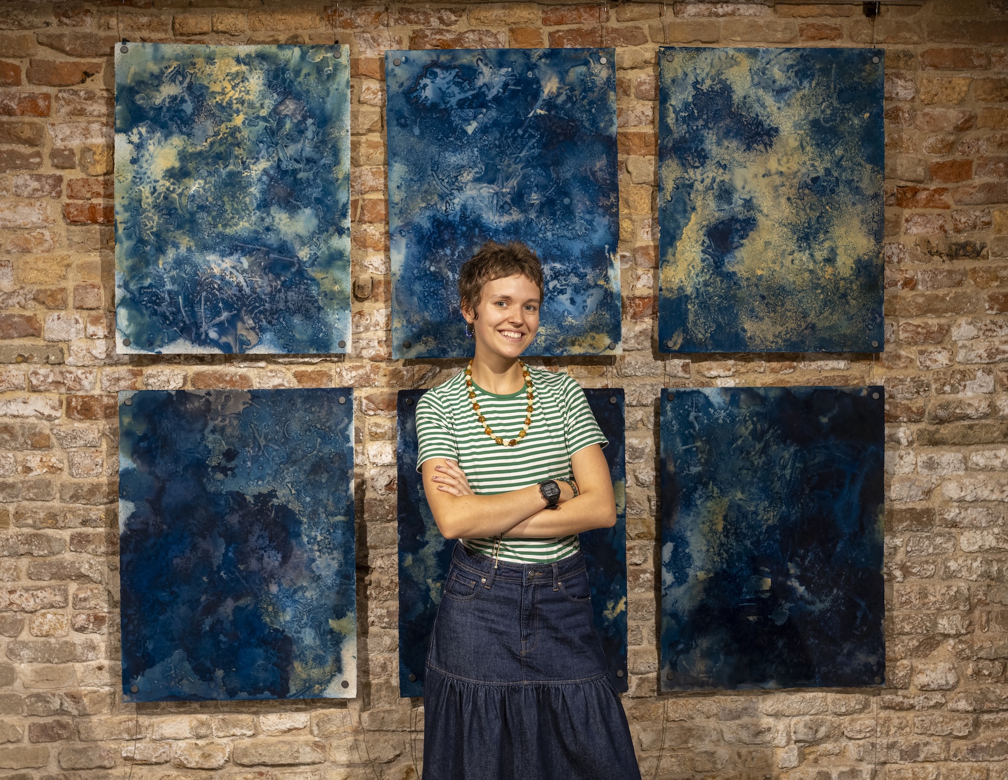 A young person stands with folded arms in front of six blue abstract paintings on a brick wall.