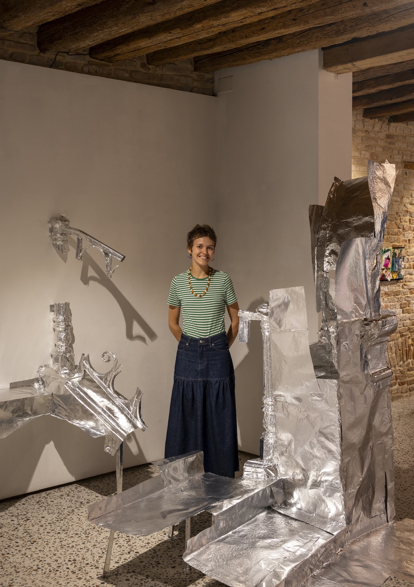 A young person in a green striped T-shirt and long dark skirt stands in a room with a wood-beamed ceiling, beside a silver foil sculptural installation.