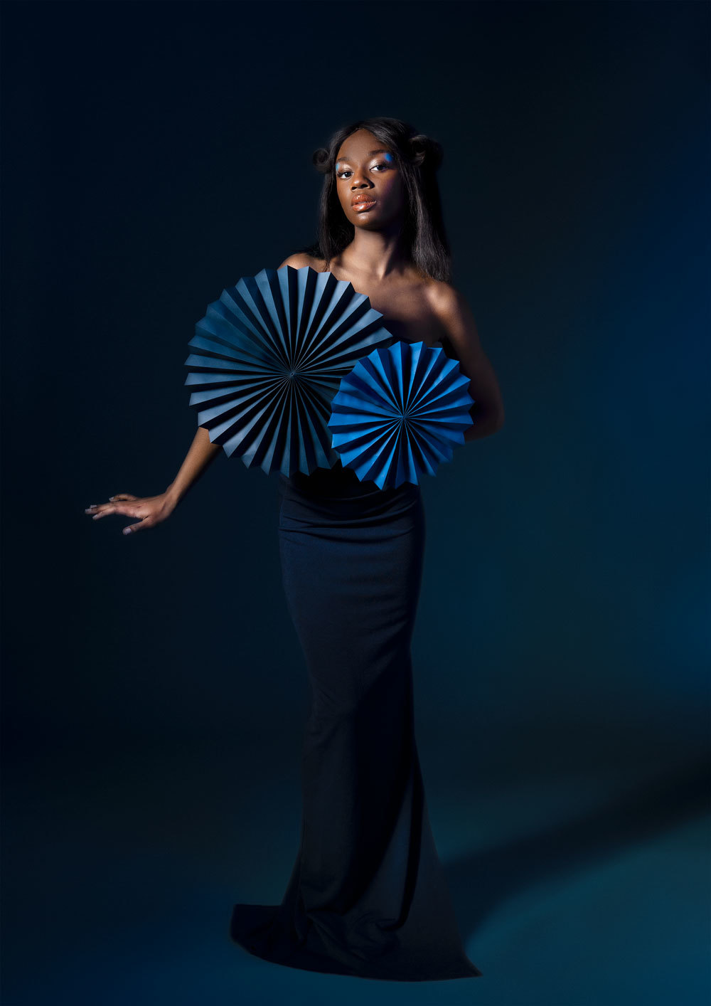 Female model in blue dress with paper fans on the dress. Taken on a blue background with dramatic lighting.