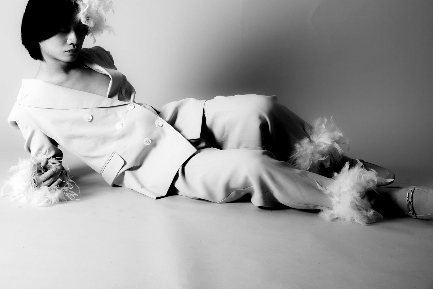 Male model lying on the floor wearing a feathered cuffed suit, in black and white.