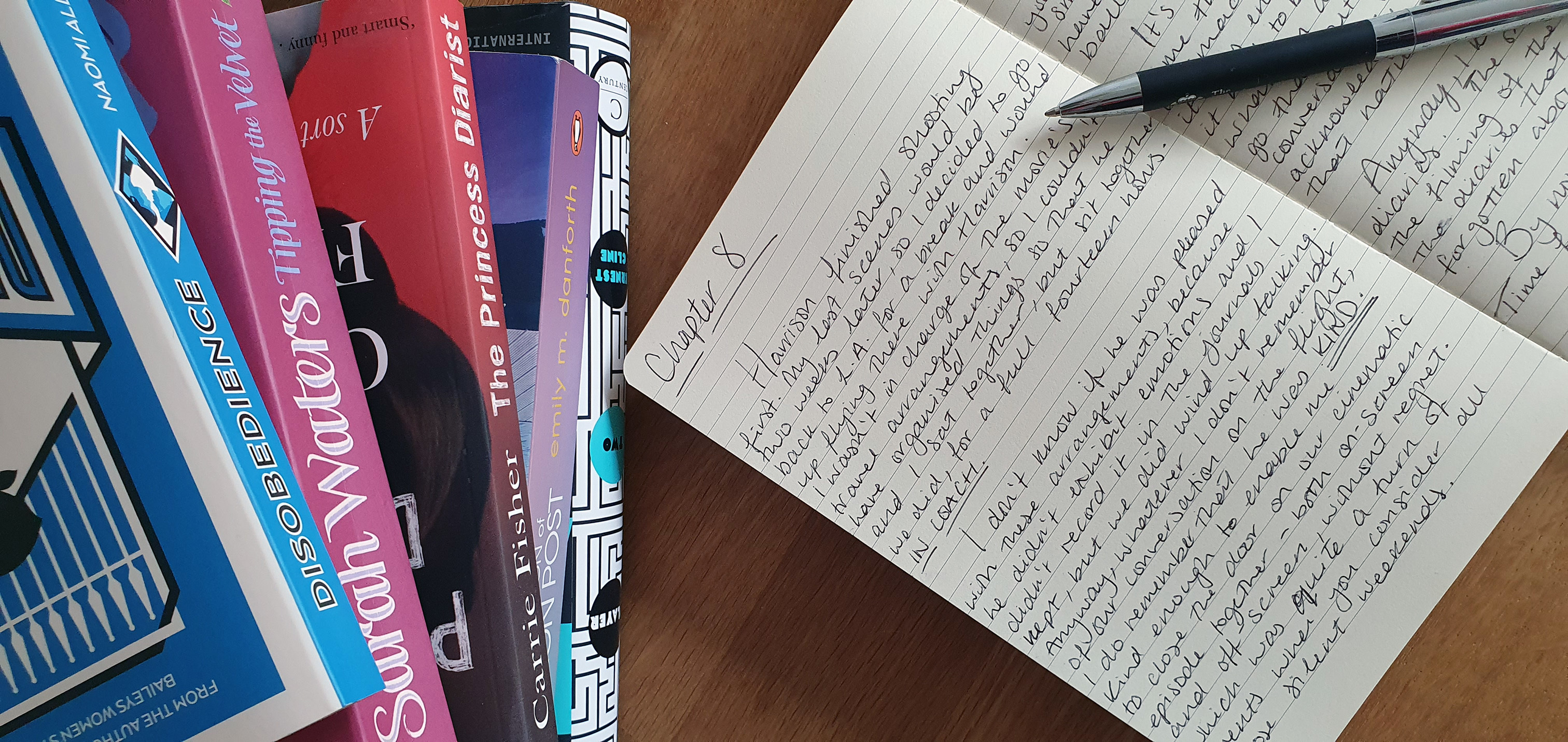 A stack of colourful books next to an open notebook. There is handwritten text in the notebook and a pen lies across the pages.
