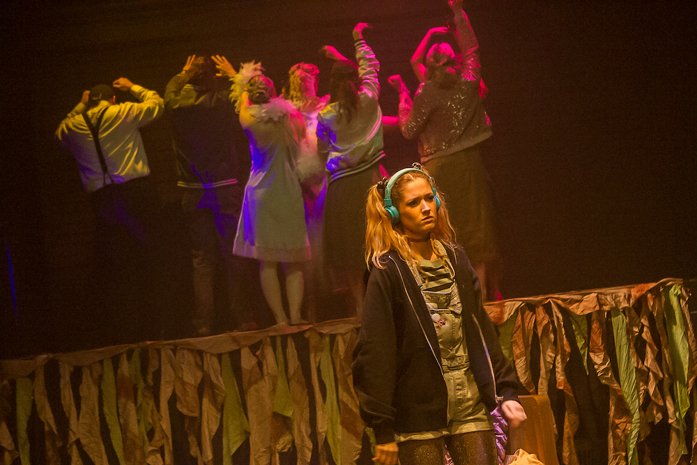 Person standing at the foot of a stage wearing headphones. A line of performers are behind them with their backs to them.
