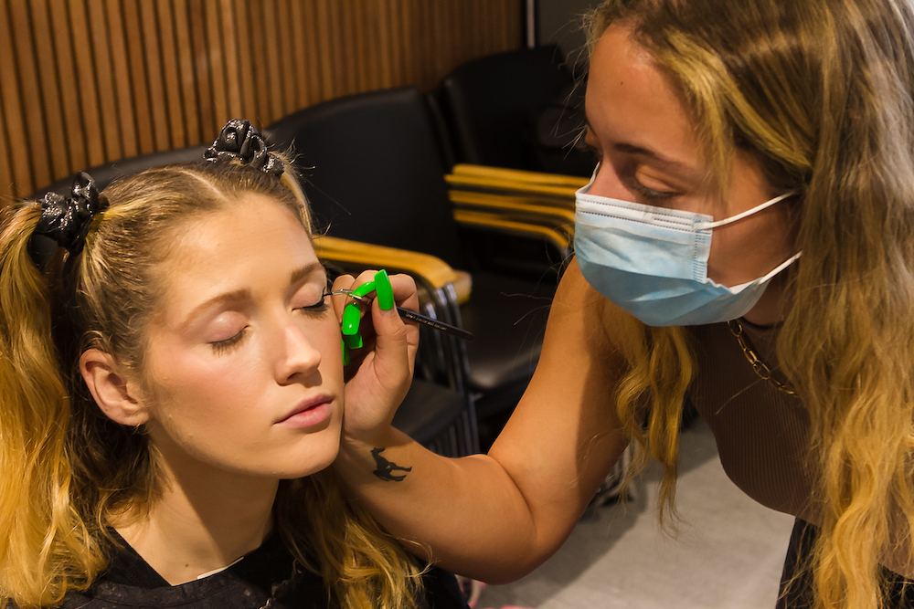 A makeup artist wearing a face mask applies makeup to an actor's face. The actor has their eyes closed.