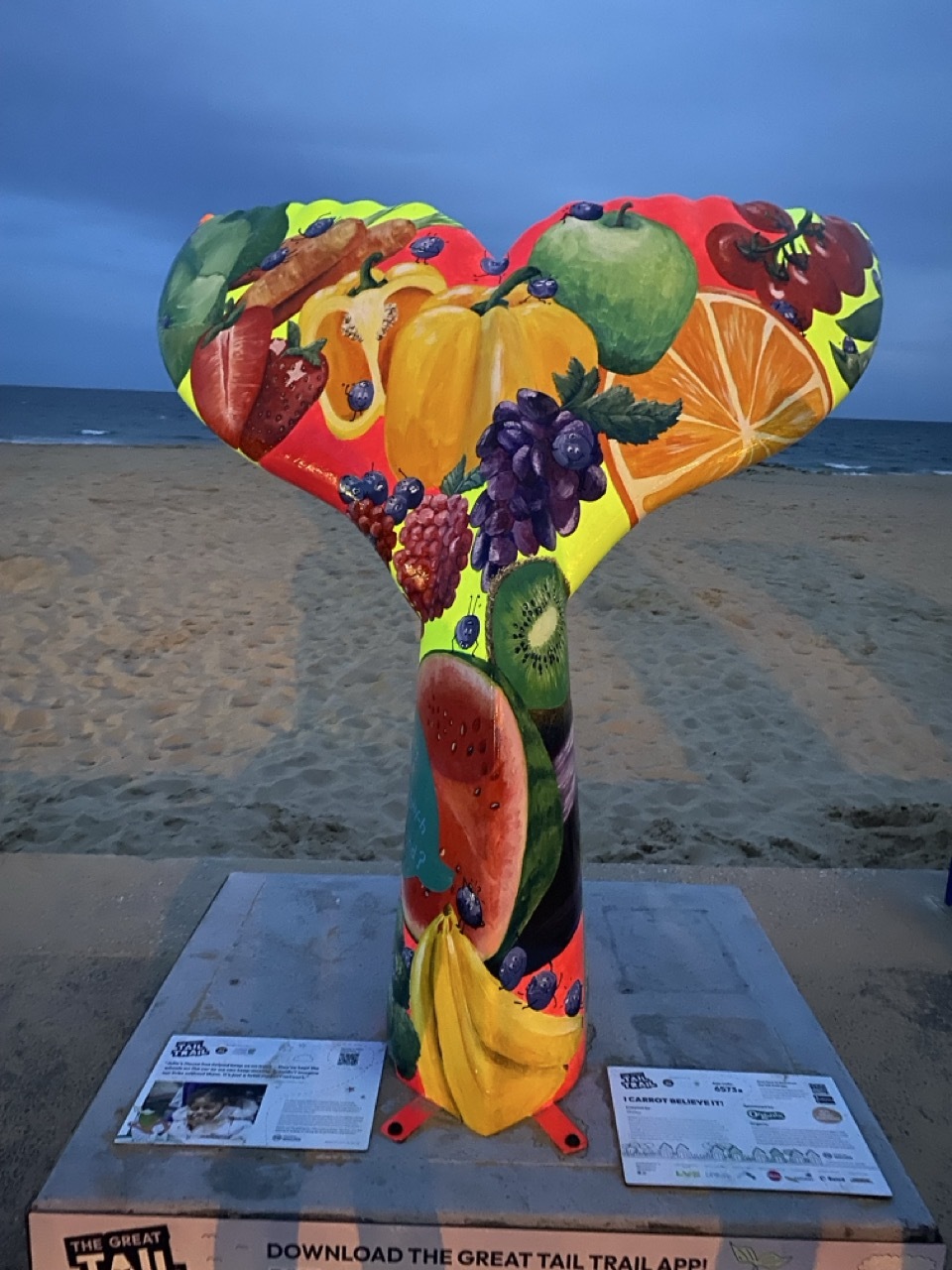 Heart-shaped sculpture covered in bright fruit imagery on beach, featuring oranges, grapes, apples in vivid colours against blue sky.