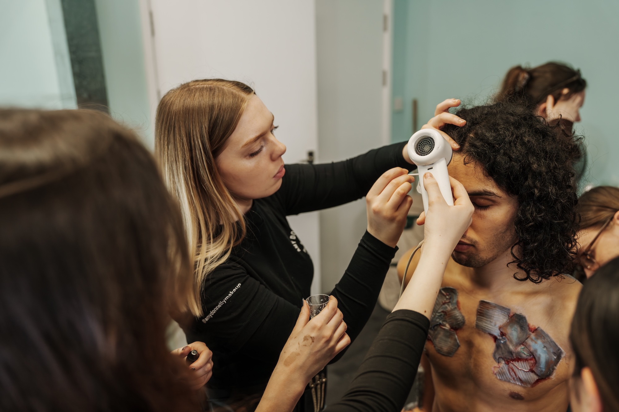 A woman with long hair applies makeup to a man with curly hair and body art, while a second woman observes in the background. The setting appears to be a makeup studio with soft lighting and muted colours on the walls.