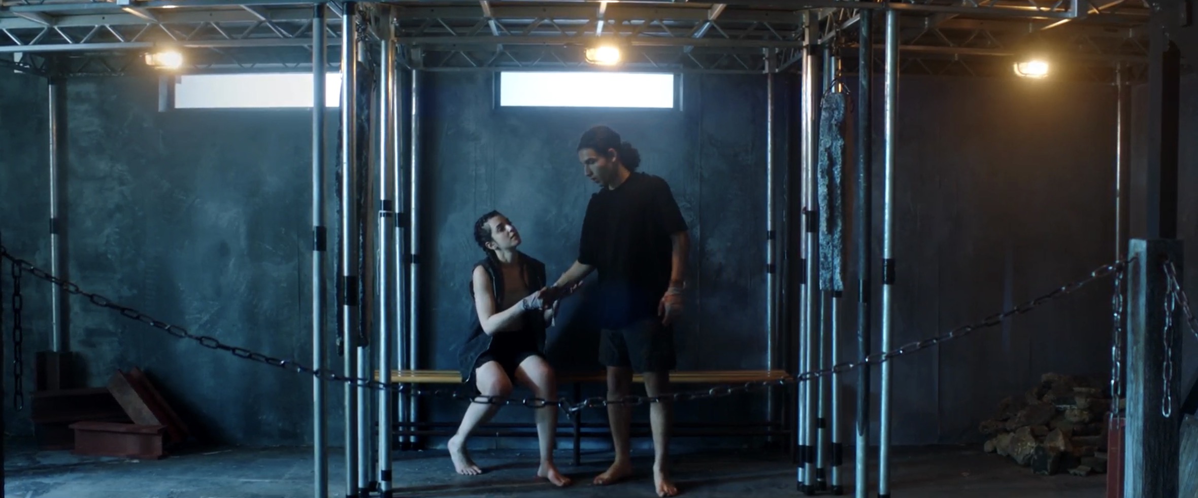 A woman in a black dress is sitting on a wooden bench, reaching out to a man standing beside her. The setting features metal structures with chains, grey walls, and dim lighting, creating an industrial atmosphere.