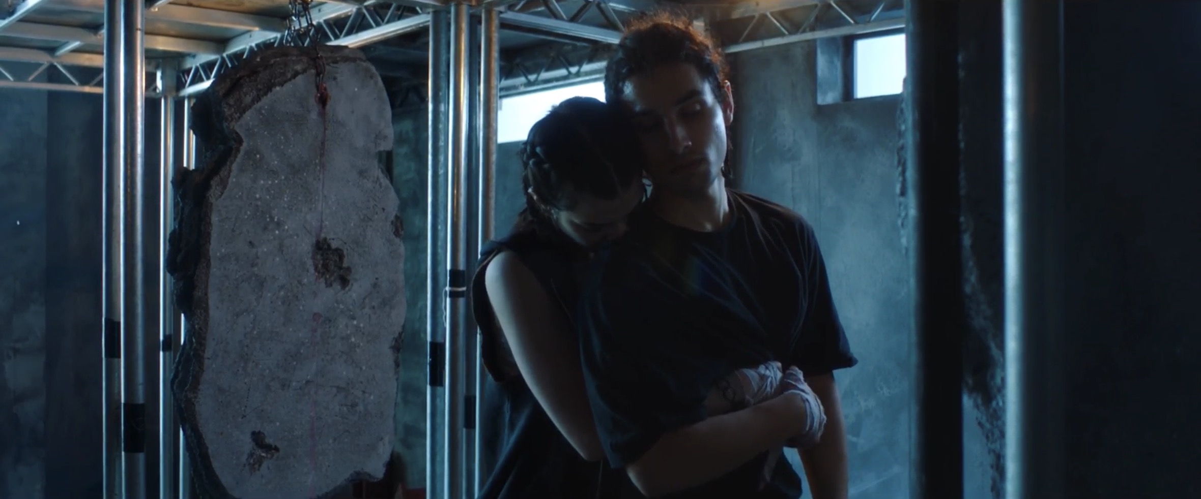 A woman with long dark hair leans her head on the shoulder of a man with short brown hair. They stand in a dimly lit room surrounded by metal supports, with a large stone slab in the background under a window.