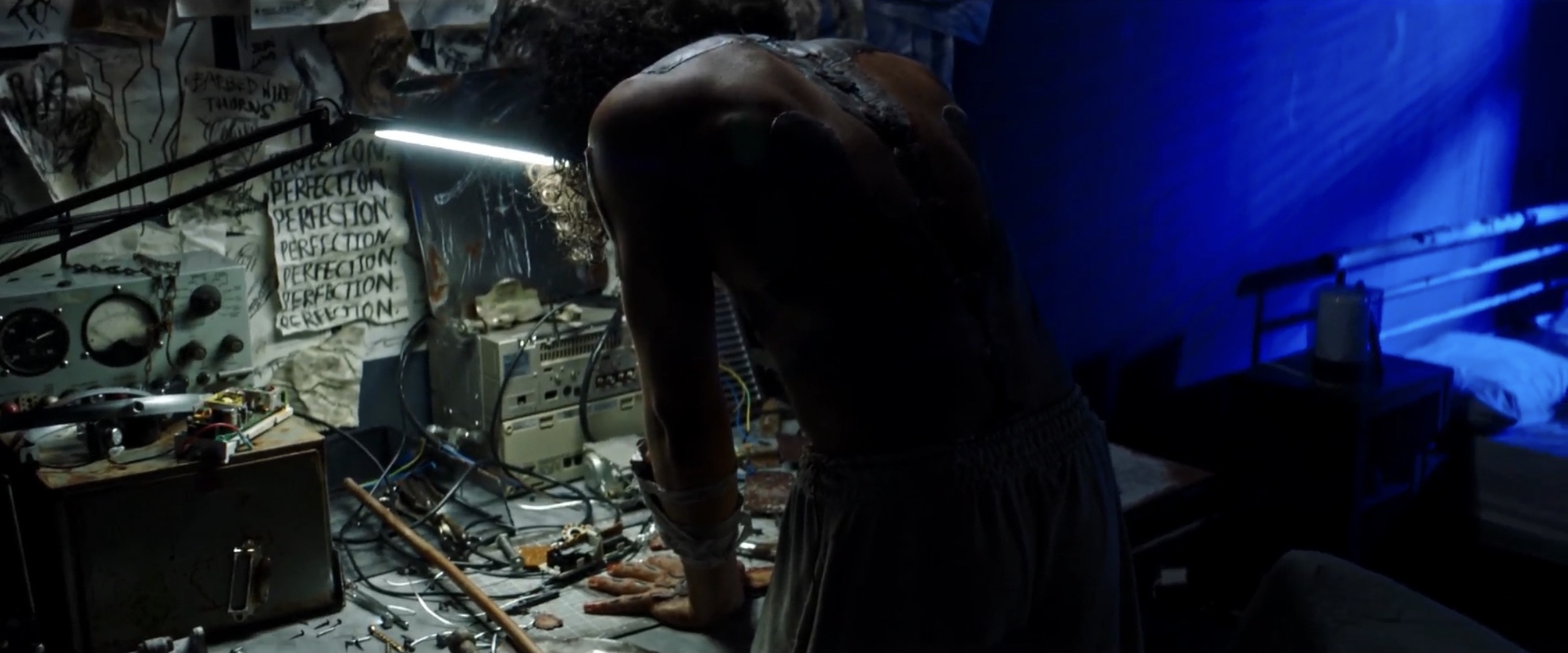 A man with a bare back leans forward over a cluttered desk filled with tools and electronic equipment. The room is dimly lit, with blue walls and a bright light illuminating the workspace.