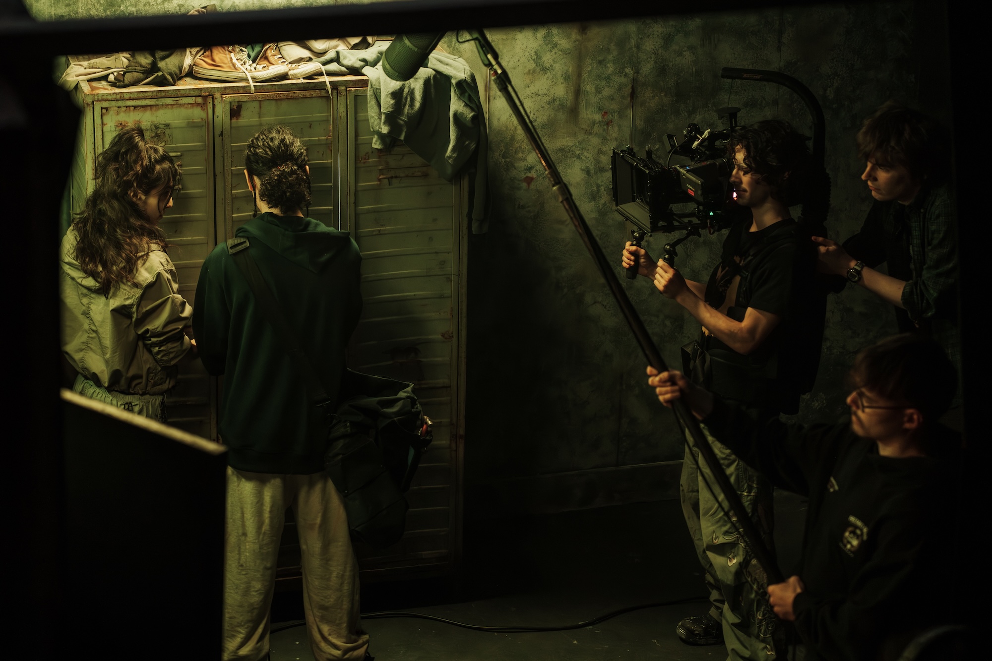 A film crew is shooting a scene in a dimly lit room. A woman in a beige jacket and a man in a black hoodie are positioned near a closet, while two crew members operate a camera and a boom microphone, capturing the action.