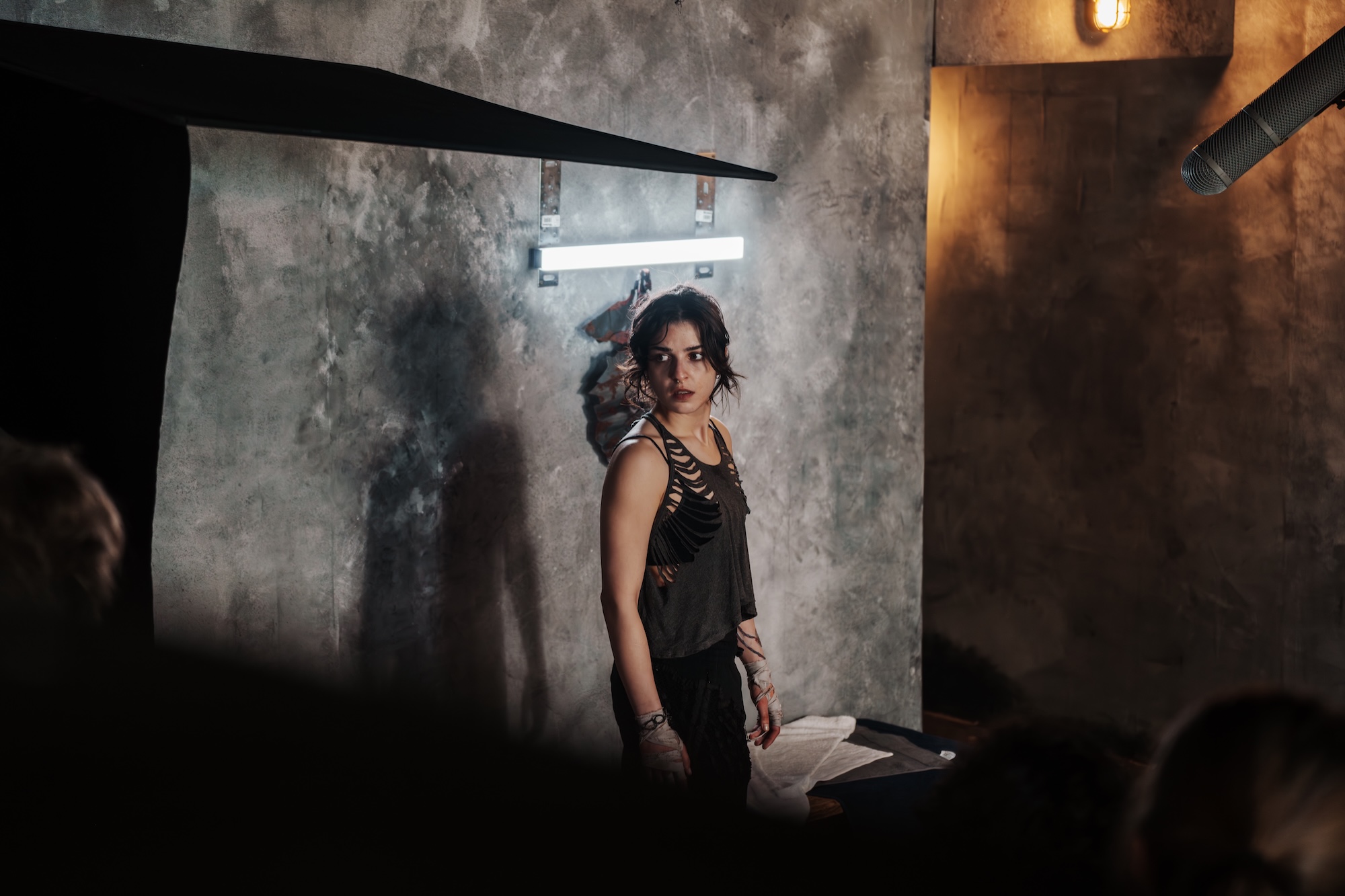 A woman stands against a textured grey wall in a dimly lit space, wearing a black tank top with embellishments. Behind her, there is a hanging light fixture and the outline of shadows on the wall, creating a contrast with the minimalistic setting.