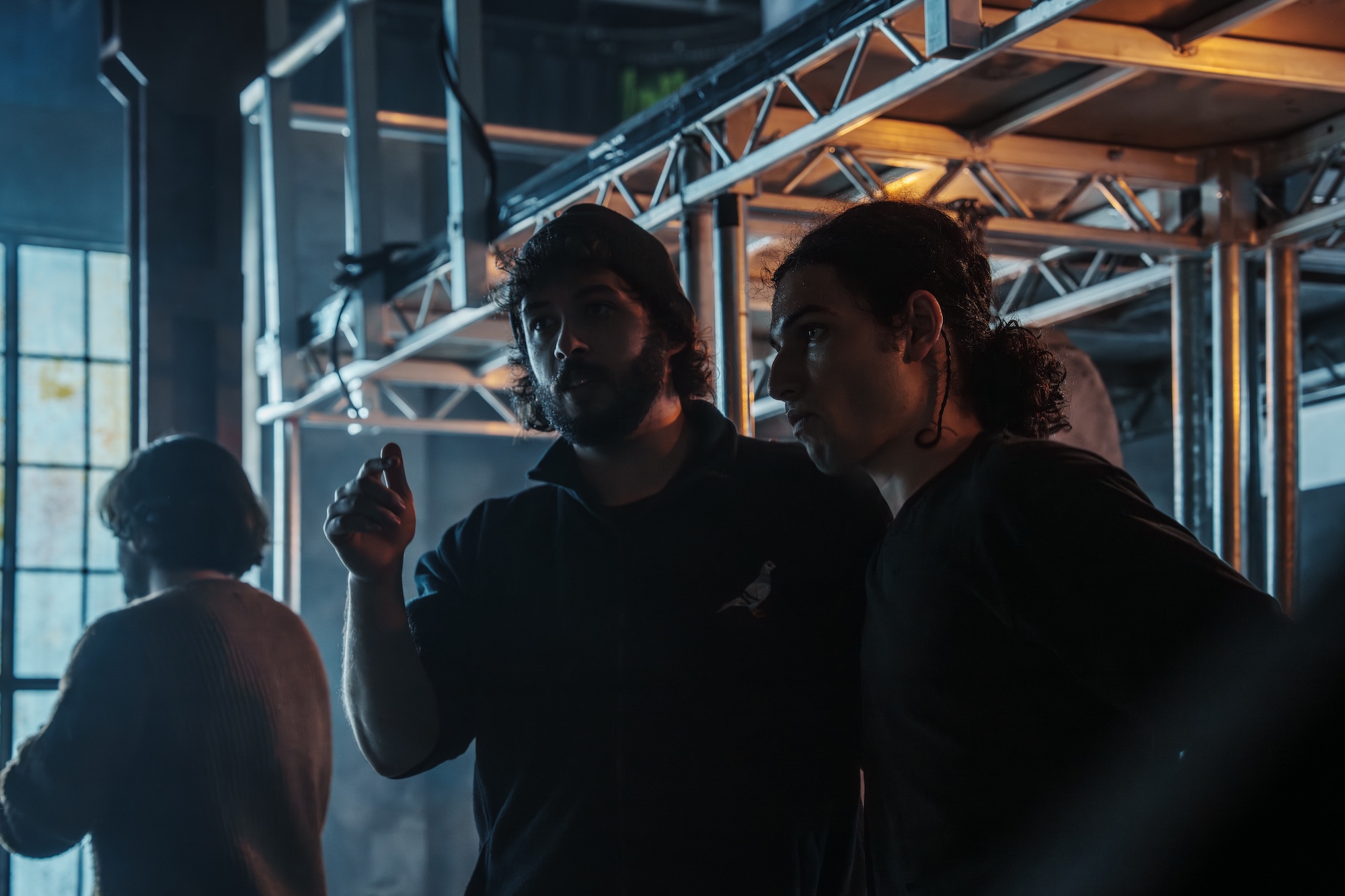 Two people are engaged in conversation in a dimly lit industrial setting. One man, wearing a dark shirt, gestures with his hand, while the other man, with a ponytail, listens attentively. In the background, additional people and metal structures are partially visible.